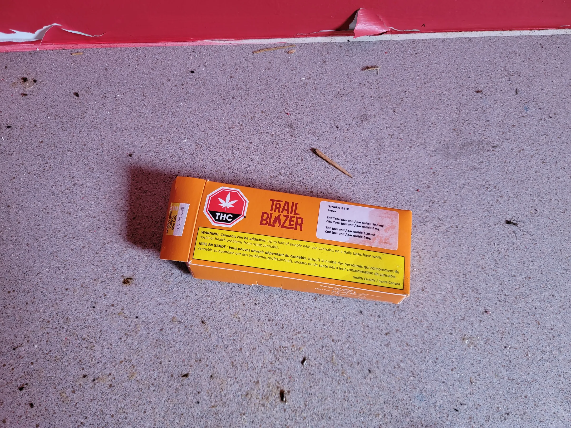 An empty cannabis pre-roll box lying on a speckled floor beside a peeling red baseboard. Litter from later visitors to the abandoned house.