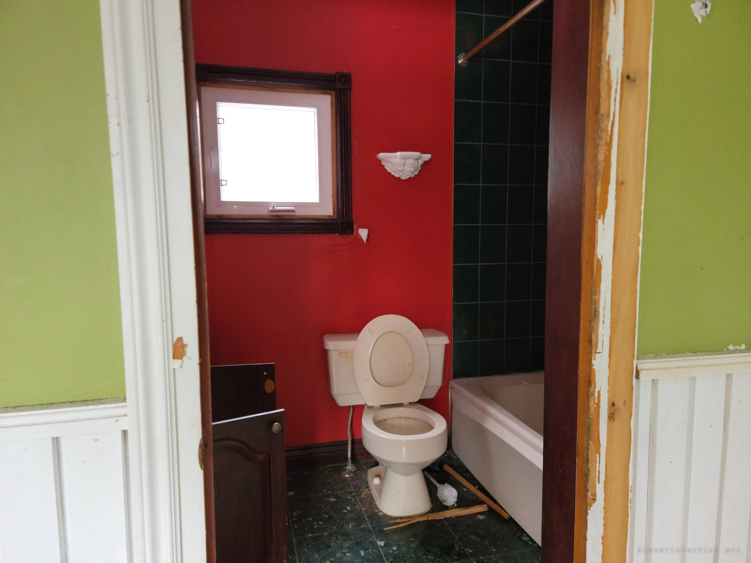 A doorway framed by green walls and white wainscoting opens onto a red bathroom with a toilet, green-tiled tub and a cherub wall shelf. Bold colours still hold in the empty house.
