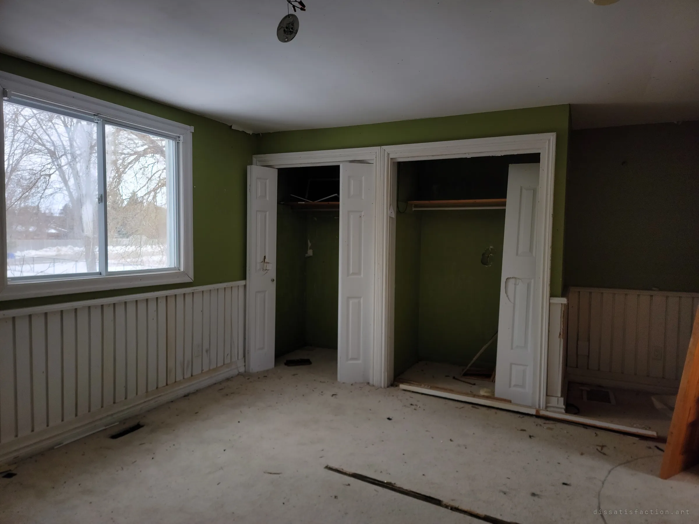 A green bedroom with white wainscoting, open bifold closets and a dirty carpet, lit by a window onto bare winter trees. The emptied closets suggest the family left long ago.