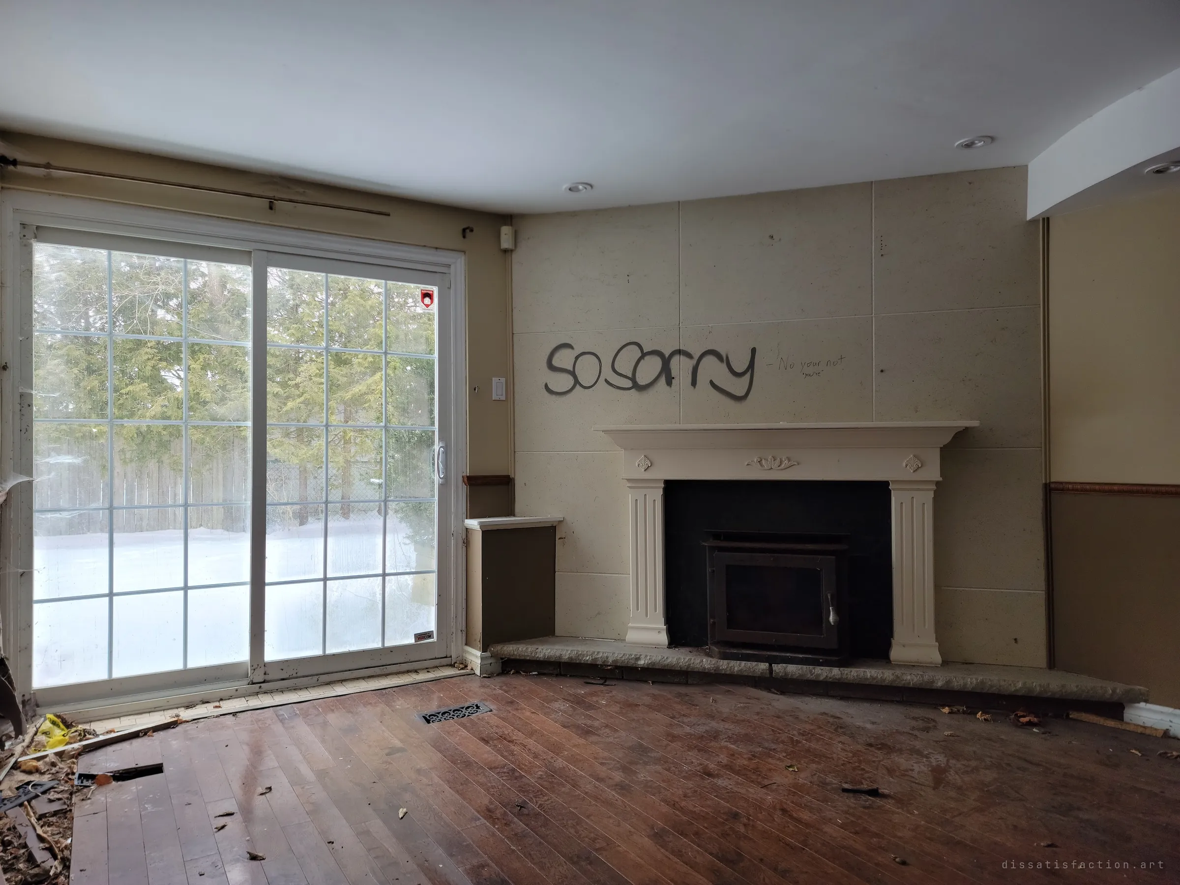 An emptied family room with a stone-panel fireplace wall sprayed with the words "so sorry", a small scribbled reply beside it, and a snowy backyard through the patio doors. The apology on the wall of an abandoned home sets the tone of the whole house.