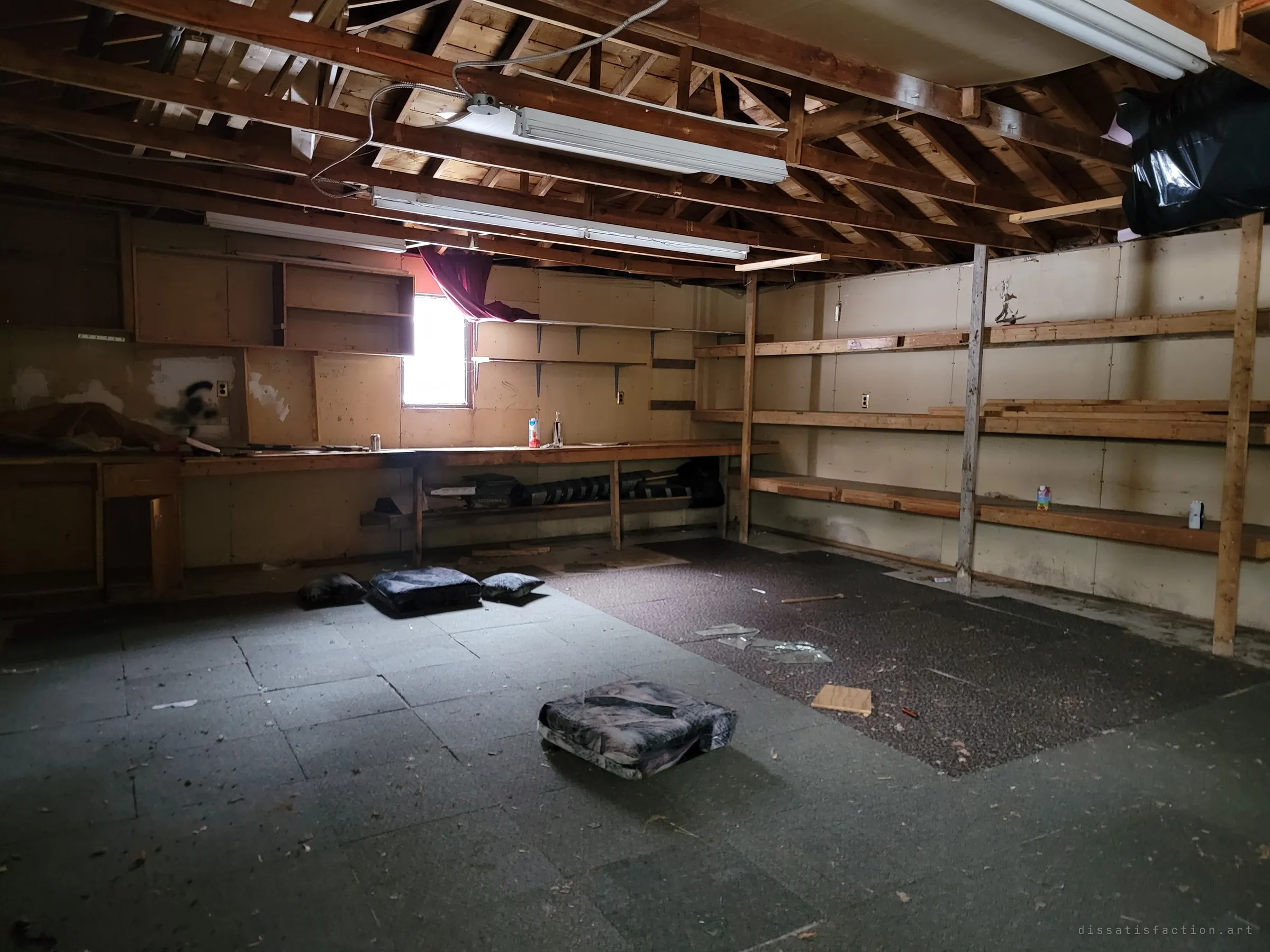 A detached garage workshop under open rafters, with long workbenches, bare shelving and cushions scattered across the tiled floor. Light from a single curtained window picks out what was left behind.