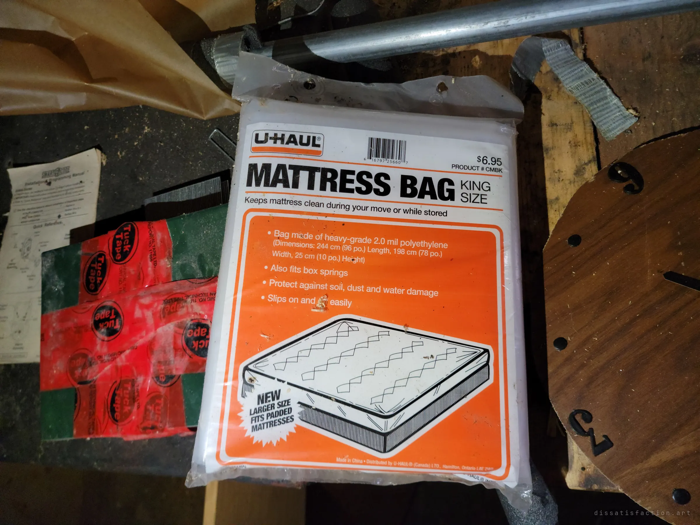An unopened king-size mattress bag lies on a workbench beside strips of red sealing tape, an old installation manual and a wooden clock face. A move that was planned but never finished.