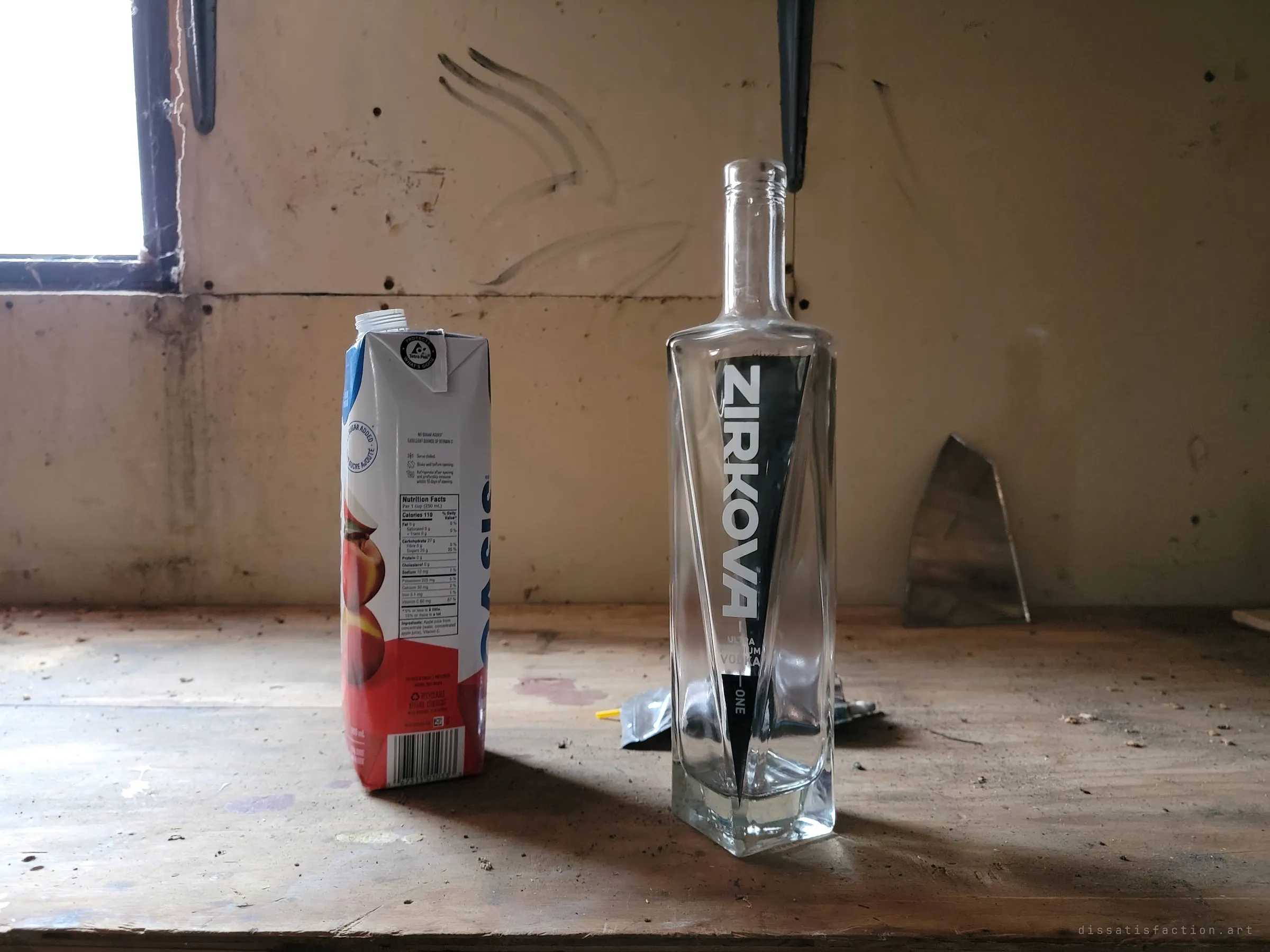 An empty vodka bottle and a juice carton stand side by side on a dusty workbench under the garage window. Leftovers from visitors who came before.