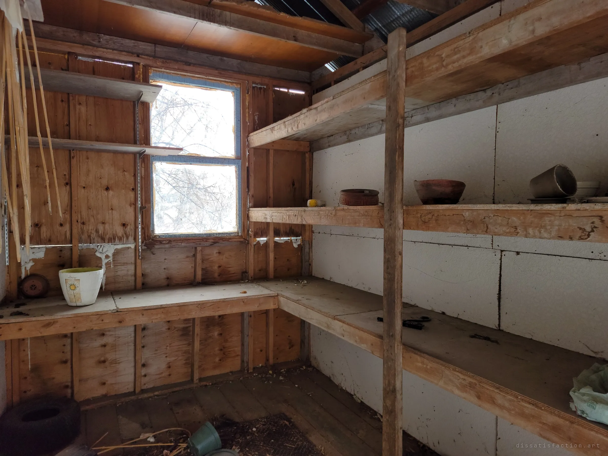 A small potting shed with plywood walls, L-shaped benches and sturdy shelves still holding a few clay pots, lit by a window full of bare branches. A garden hobby frozen mid-season.