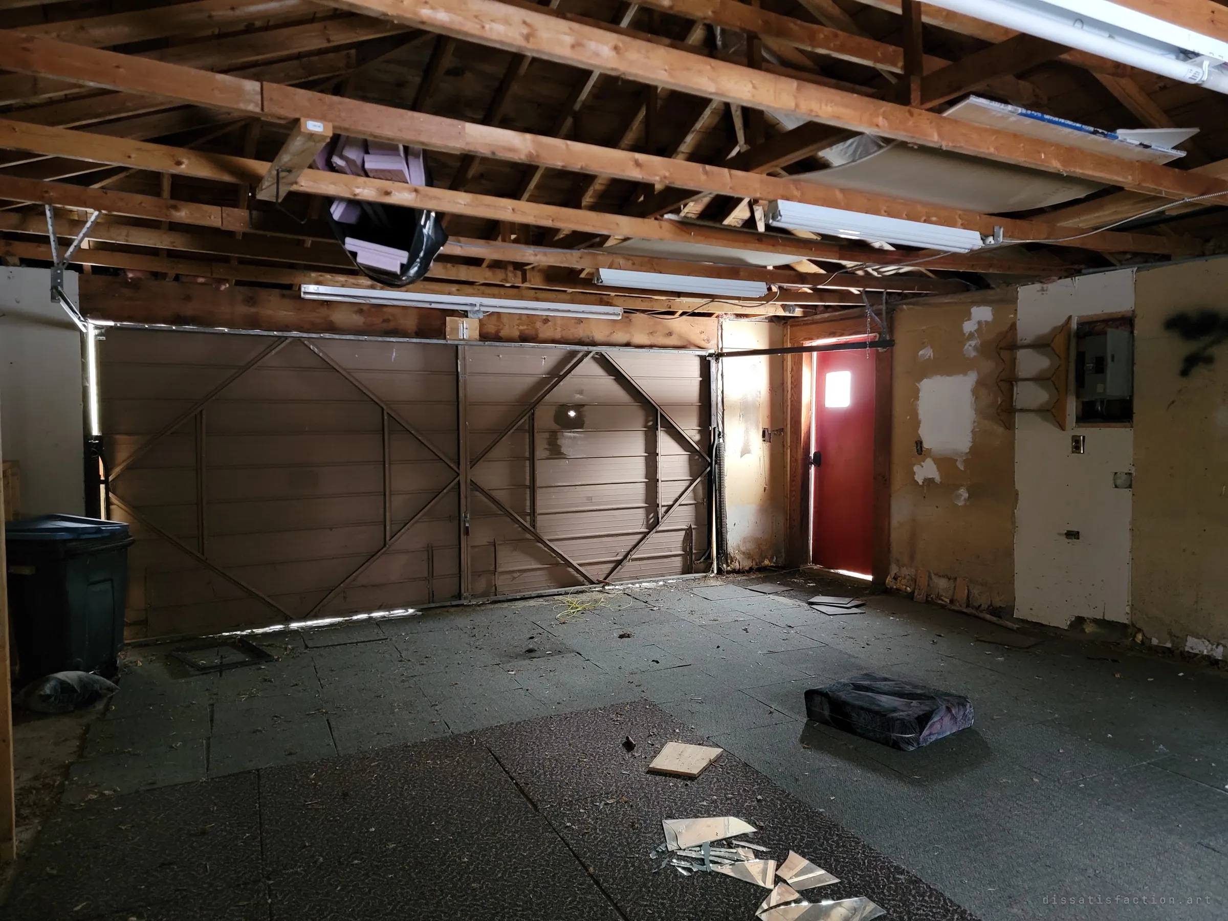 The double garage seen from inside, facing its closed brown door and a red side door glowing with daylight, with broken mirror shards on the floor. Open rafters and hanging fluorescent tubes frame the empty space.