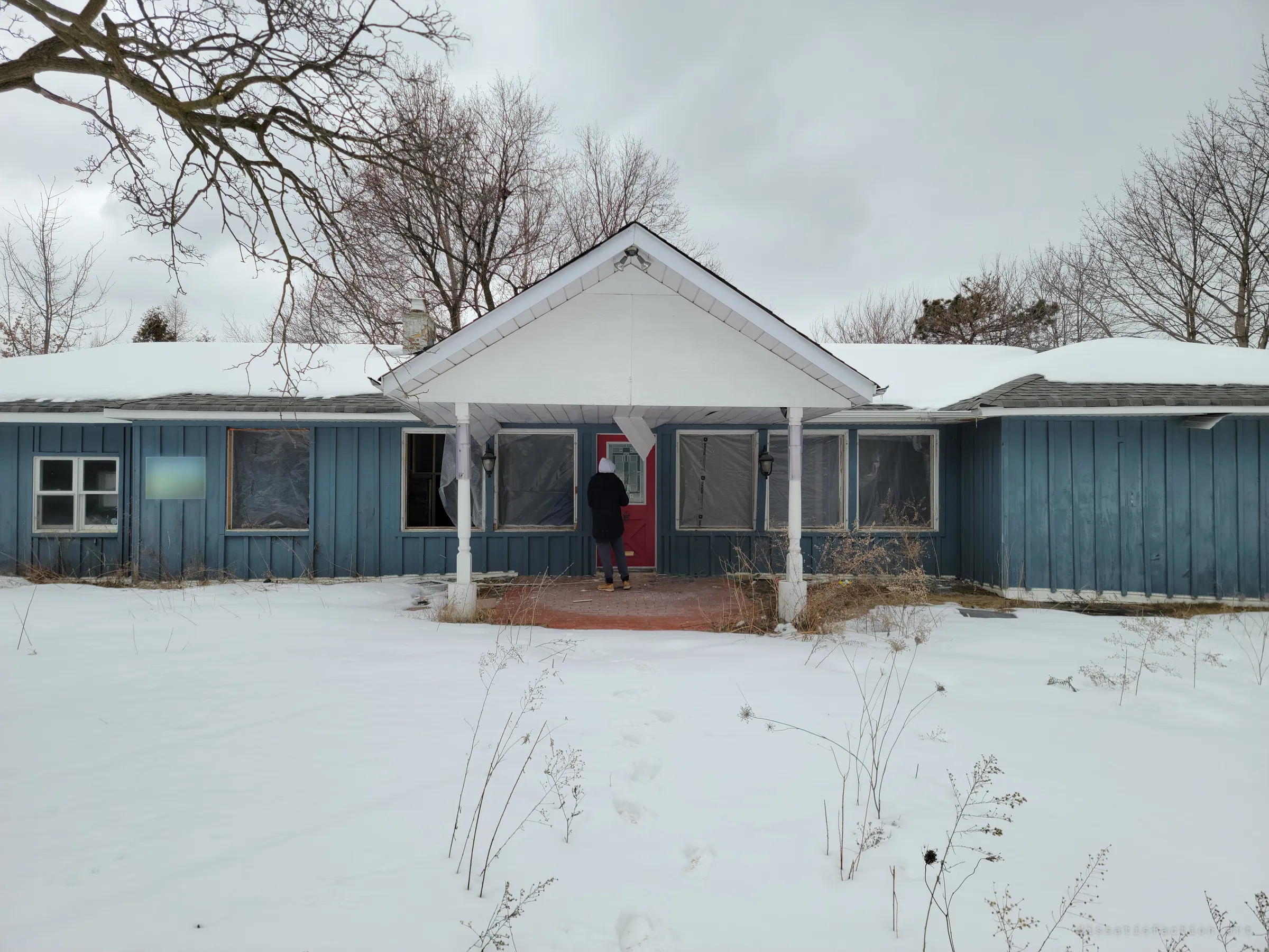 A snow-covered blue single-storey building with a white gabled porch, its windows sealed in plastic, while a hooded figure waits at the red front door. The empty yard and wrapped windows show a building mid-renovation and closed to its users.