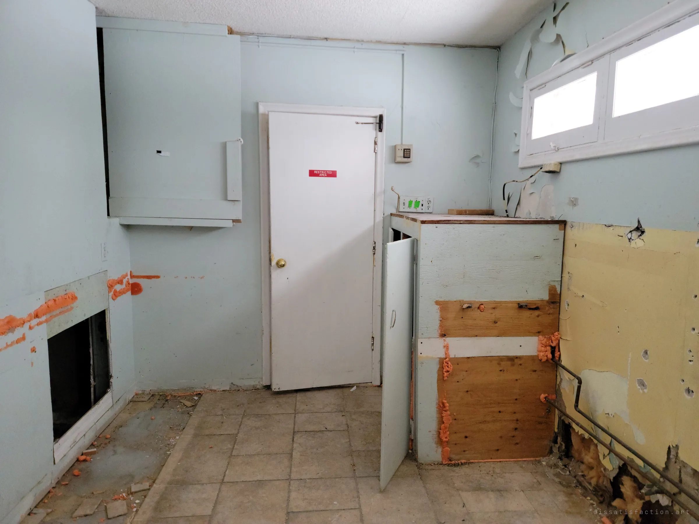 A stripped utility room with a restricted-area door, a keypad on the wall and orange foam sealing torn-open panels. The gutted cabinets and exposed pipes show the interior being taken apart.
