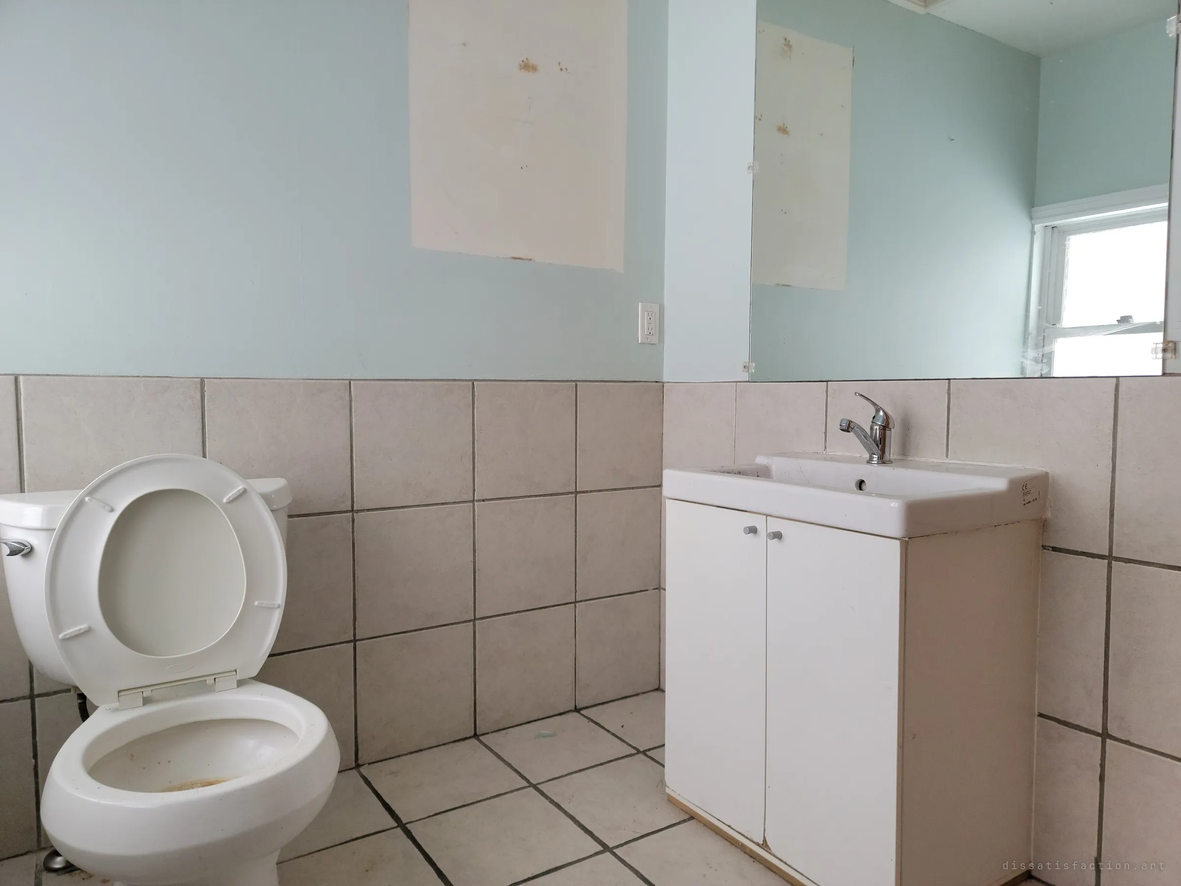 A small washroom with a stained toilet, a child-height vanity and a large mirror, with a pale patch where a poster once hung. It is a quiet trace of the young children who used this space.