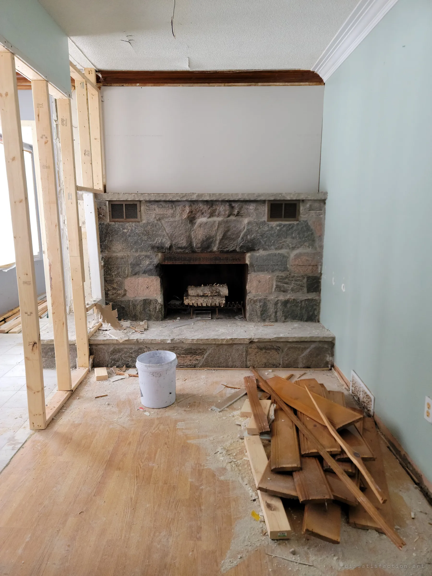 A fieldstone fireplace stands behind new wall studs, with a paint bucket and a pile of torn-out wood panelling on the floor. The old hearth contrasts with the raw framing of the renovation.