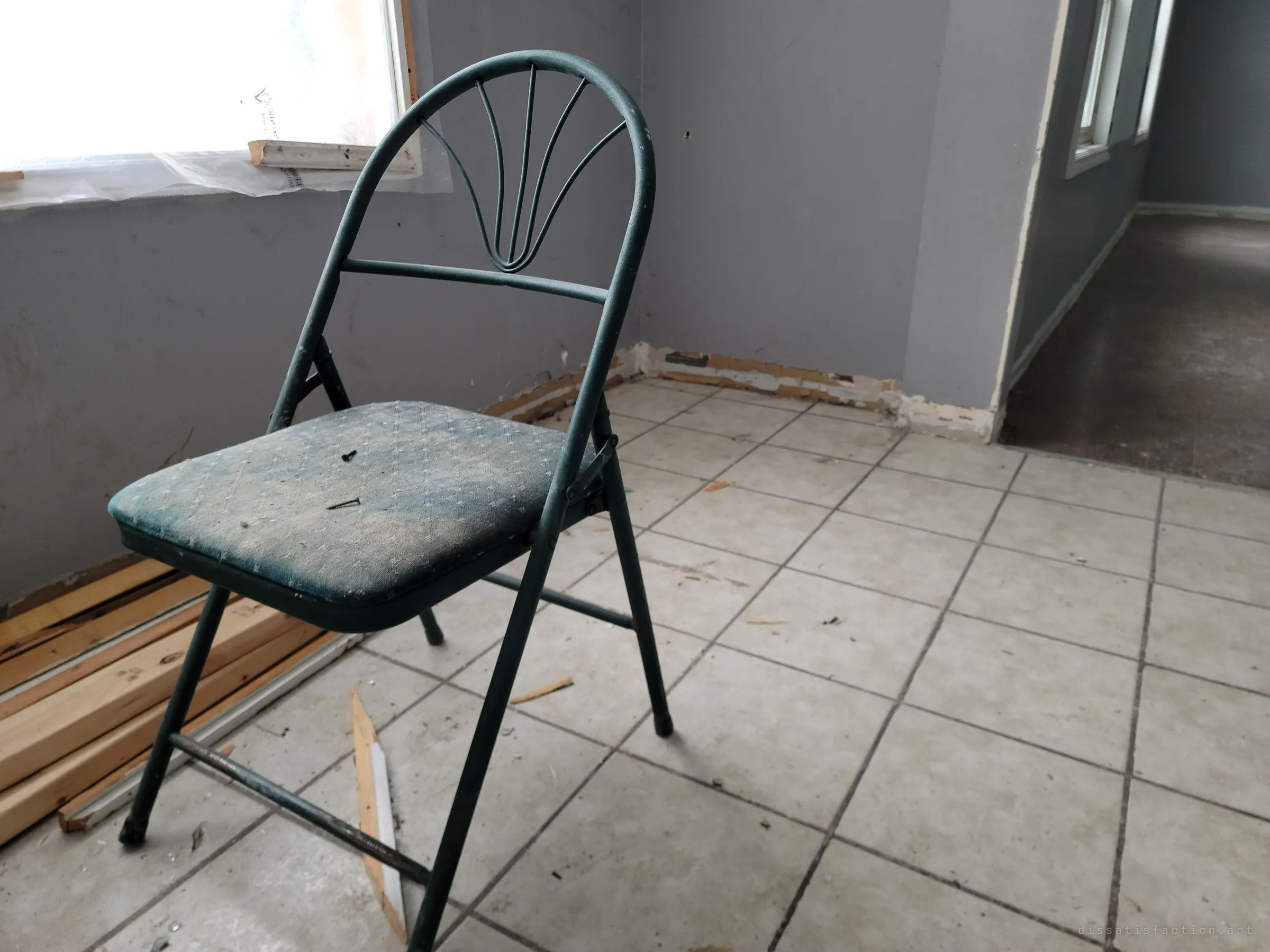 A dusty green folding chair sits alone on a tiled floor by a plastic-covered window, with stripped baseboards and scrap lumber around it. A single abandoned seat in an emptied room.