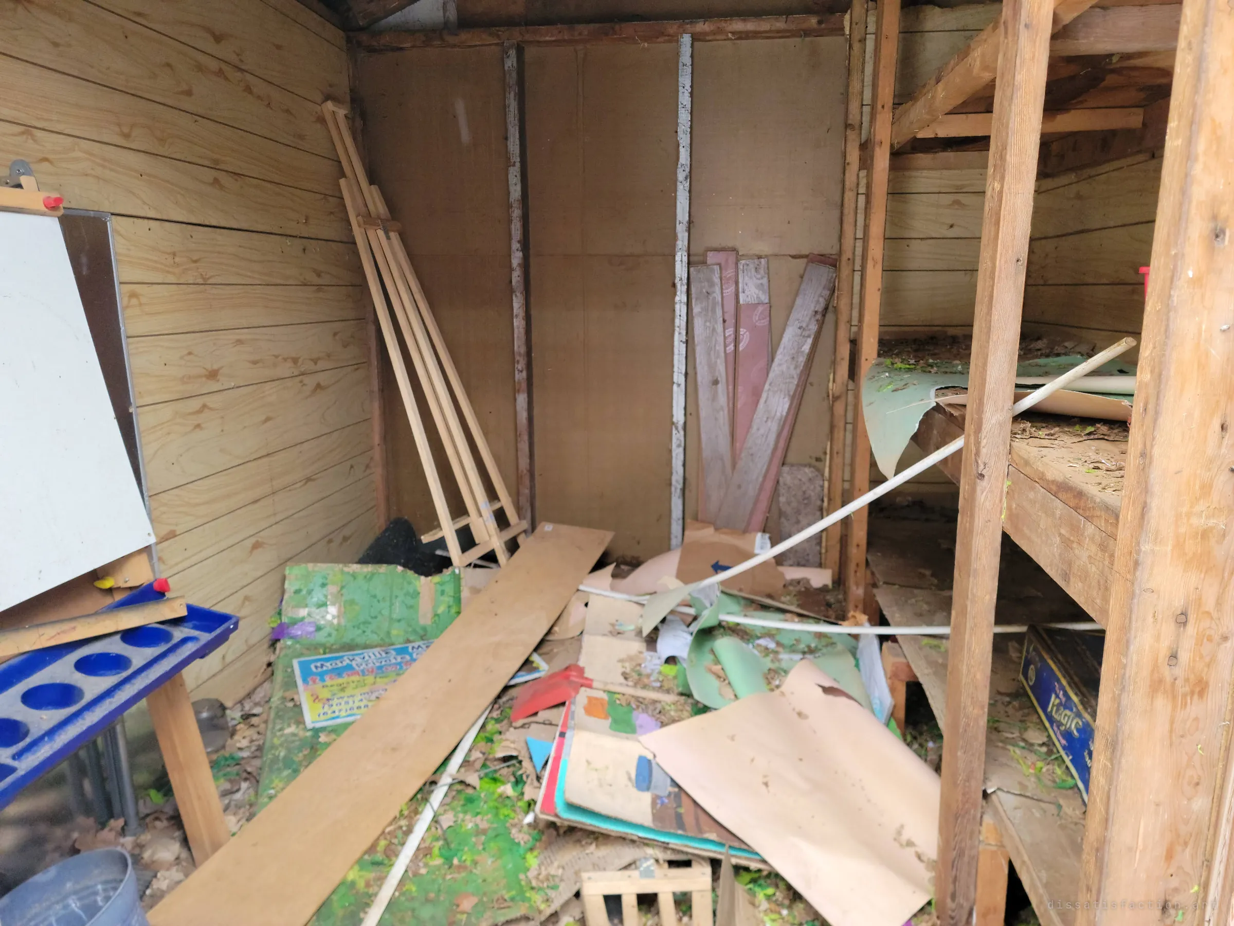 A wooden shed full of cast-off school things: an easel, a paint tray, rolled paper, boards and a discarded sign among the leaves. The heap shows the classroom's contents being cleared out.
