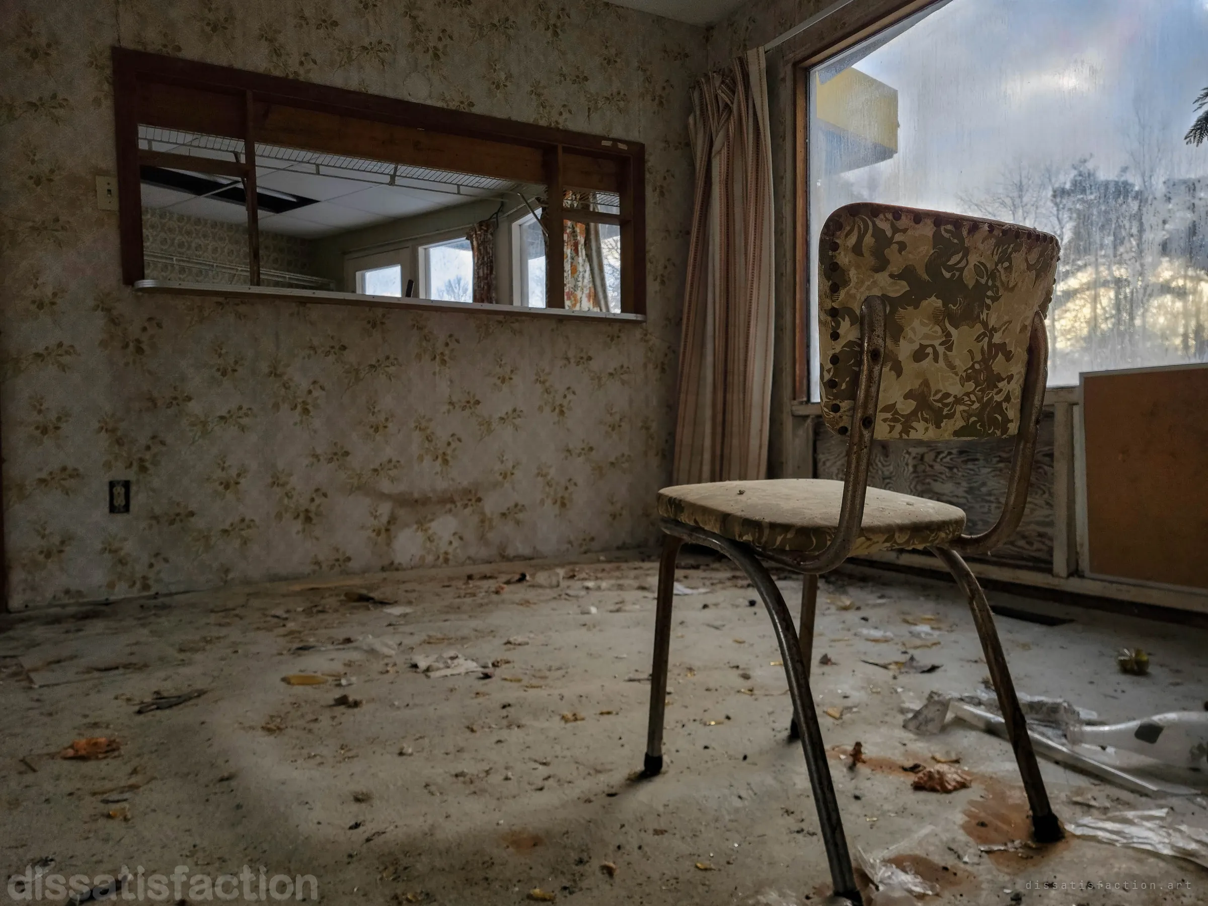 A lone chrome-legged kitchen chair with floral vinyl sits on a debris-strewn floor beside a frosted picture window, a pass-through opening in the patterned wallpaper looking into the next room. The single chair gives the empty house a waiting, staged feel.