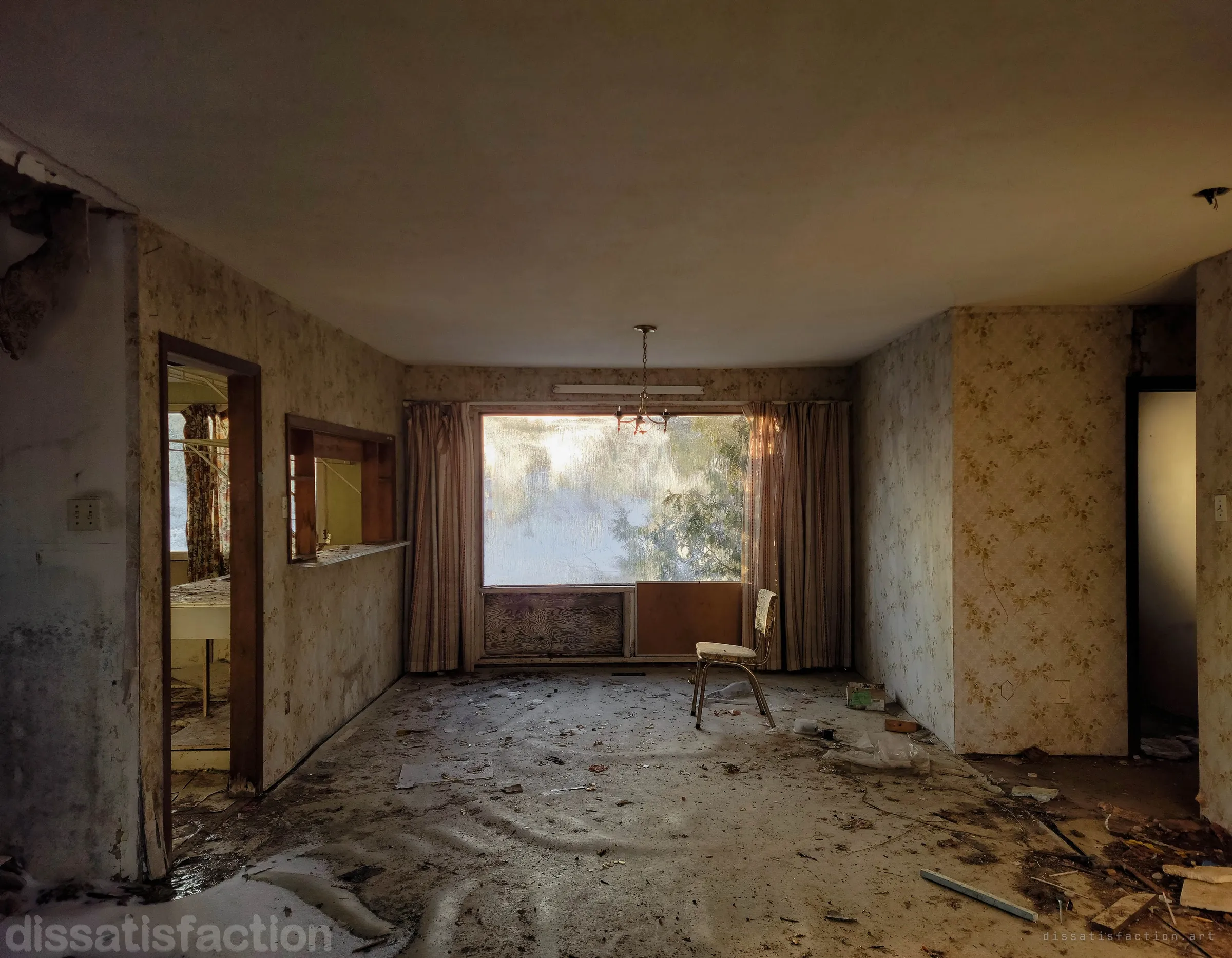 Wide view of a stripped living and dining room: floral wallpaper, a small chandelier and one chair silhouetted against a large curtained window, with a kitchen pass-through and doorways on either side. It shows the whole layout of the abandoned model interior in low winter light.