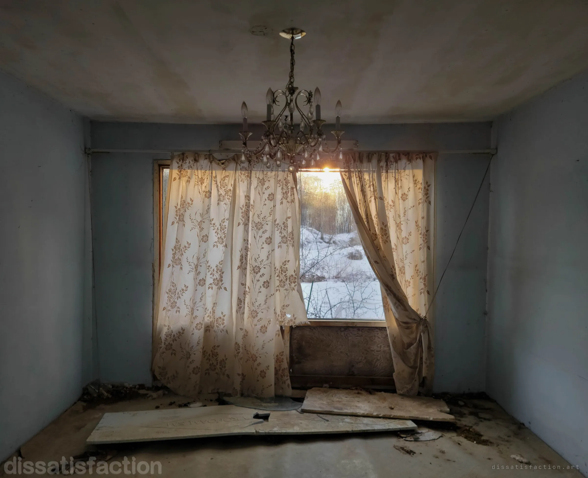 A crystal chandelier hangs in a pale blue bedroom in front of torn floral curtains, the low sun flaring through a gap onto a snowy slope outside. Broken boards lie on the bare floor below.