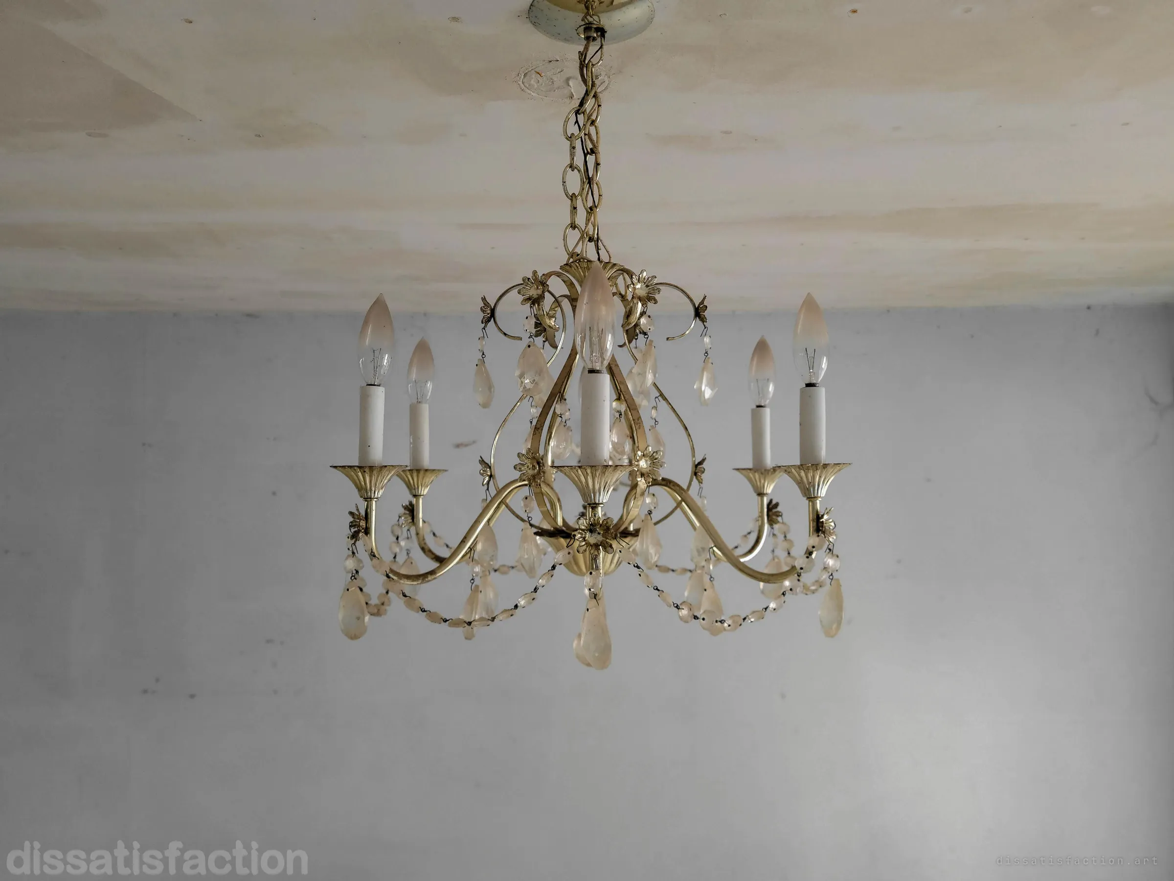 Close study of a brass chandelier with candle bulbs and hanging crystal drops against a stained ceiling and grey wall. A small remnant of the home's show-piece finish that survived the decay.