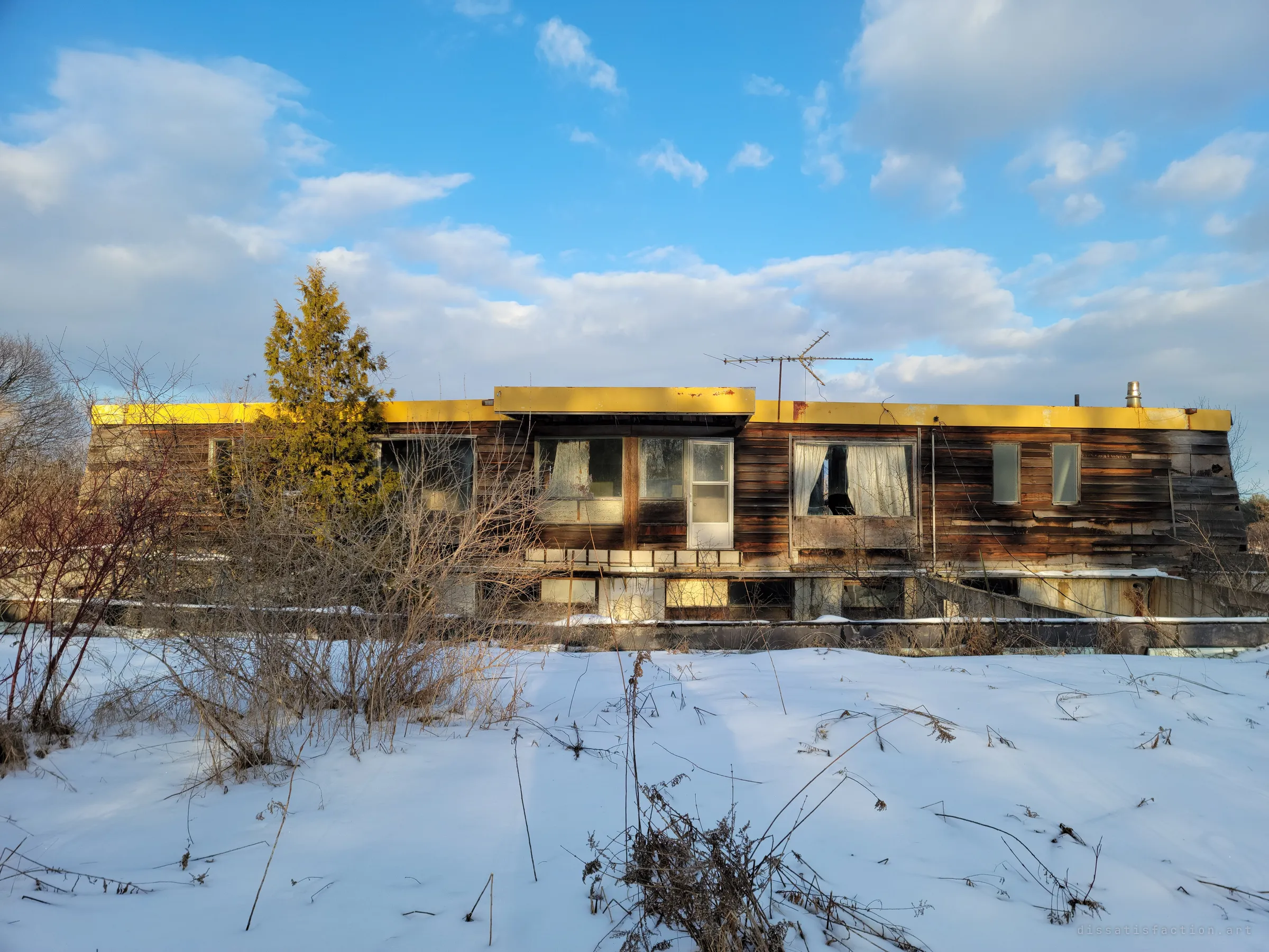 The house from across a snowy field: a weathered wood-clad upper floor under a flat roof trimmed in faded yellow, perched on a concrete base, with an old TV antenna against a blue sky.