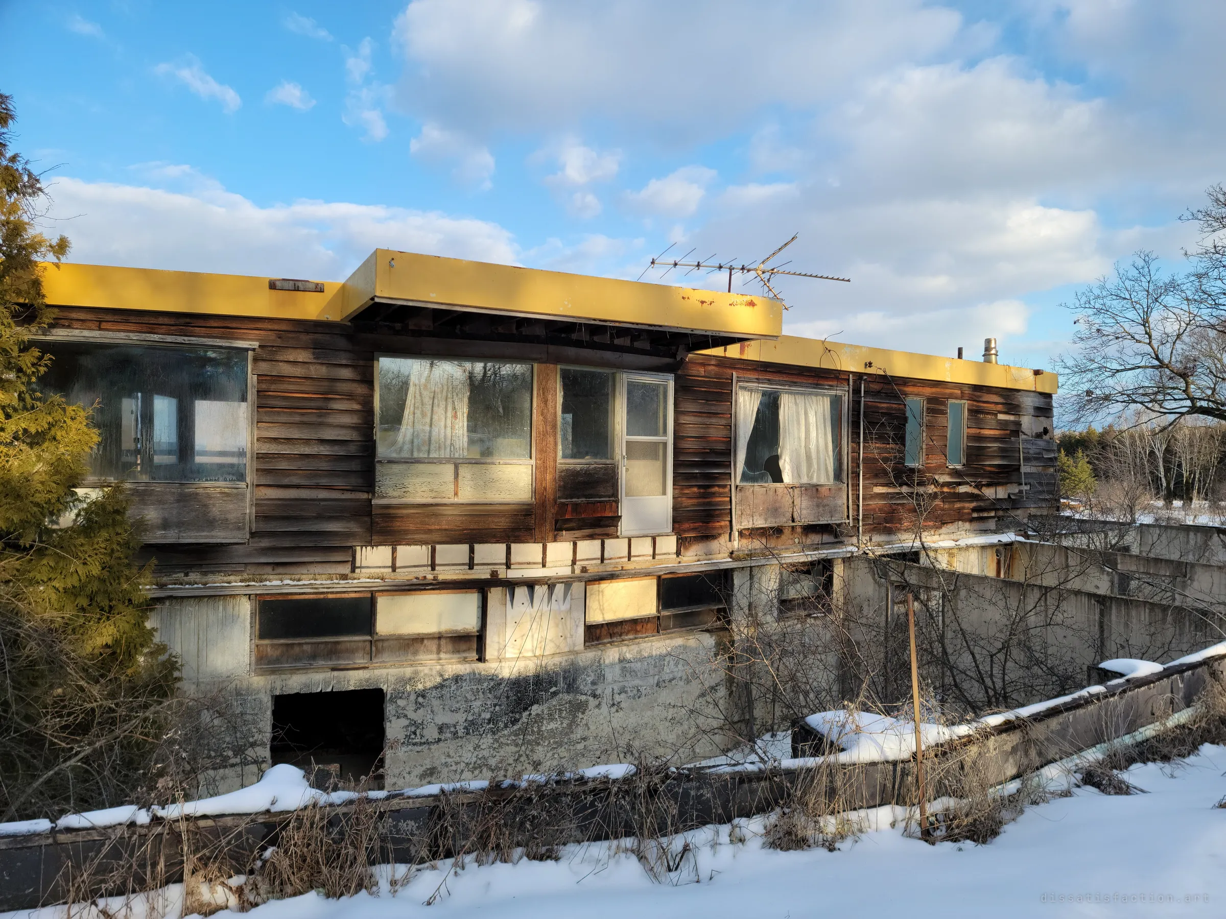 Three-quarter view of the facade: peeling cedar siding, curtained windows and a white door on the cantilevered upper floor, with a boarded concrete basement level and snow-covered retaining walls below.