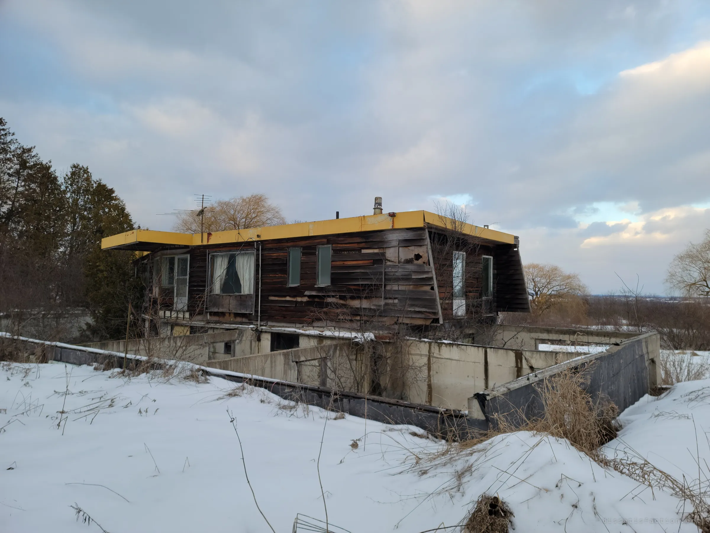 The house seen from a snowy rise under heavy dusk cloud, its yellow-trimmed roof floating over a maze of concrete foundation walls. The unfinished walls hint at plans that were never completed.