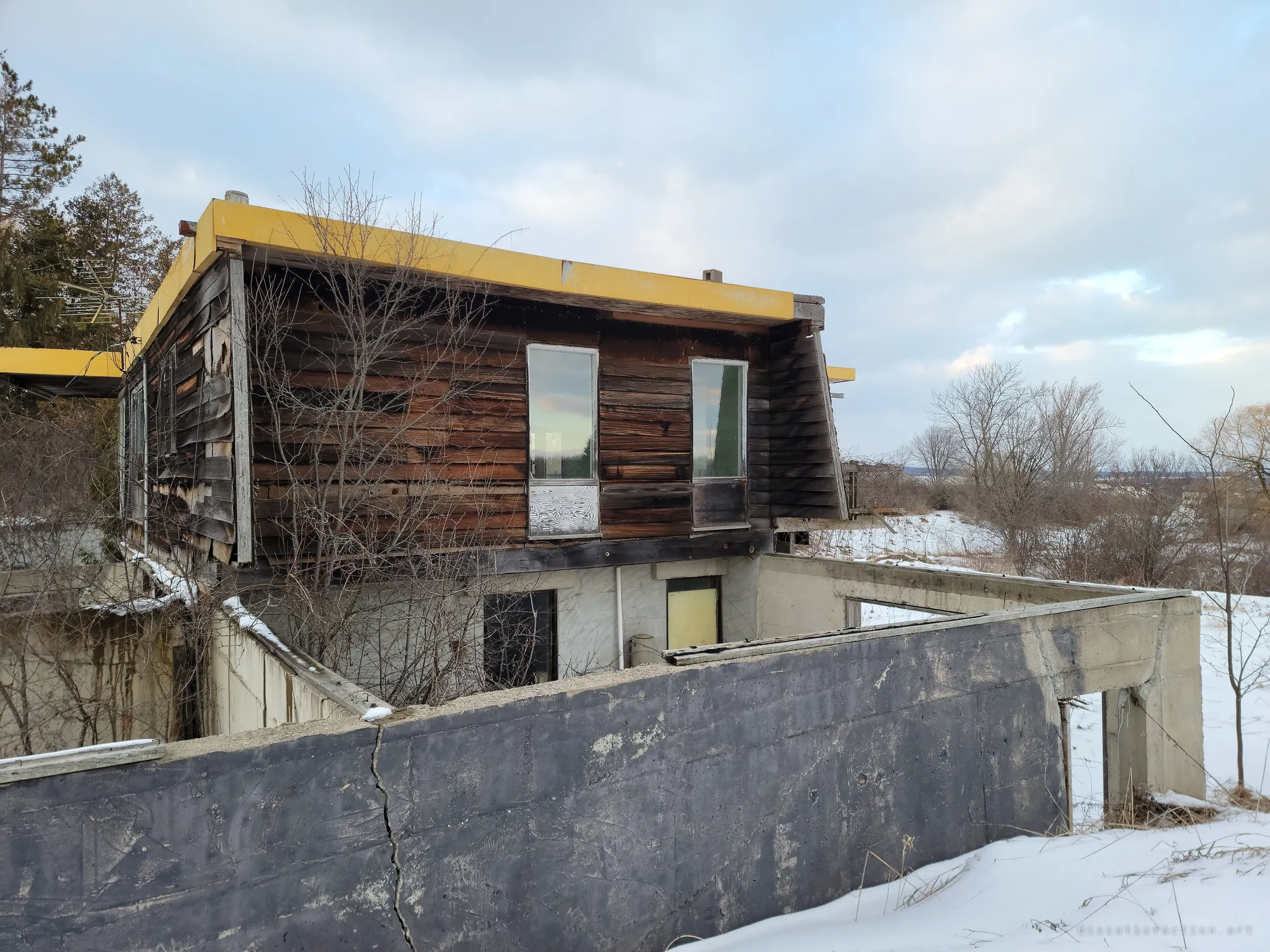 The end wall of the house overhangs a cracked, tar-blackened concrete wall, with bare trees and snowy fields falling away behind. It shows how the timber box sits on top of the unfinished concrete shell.