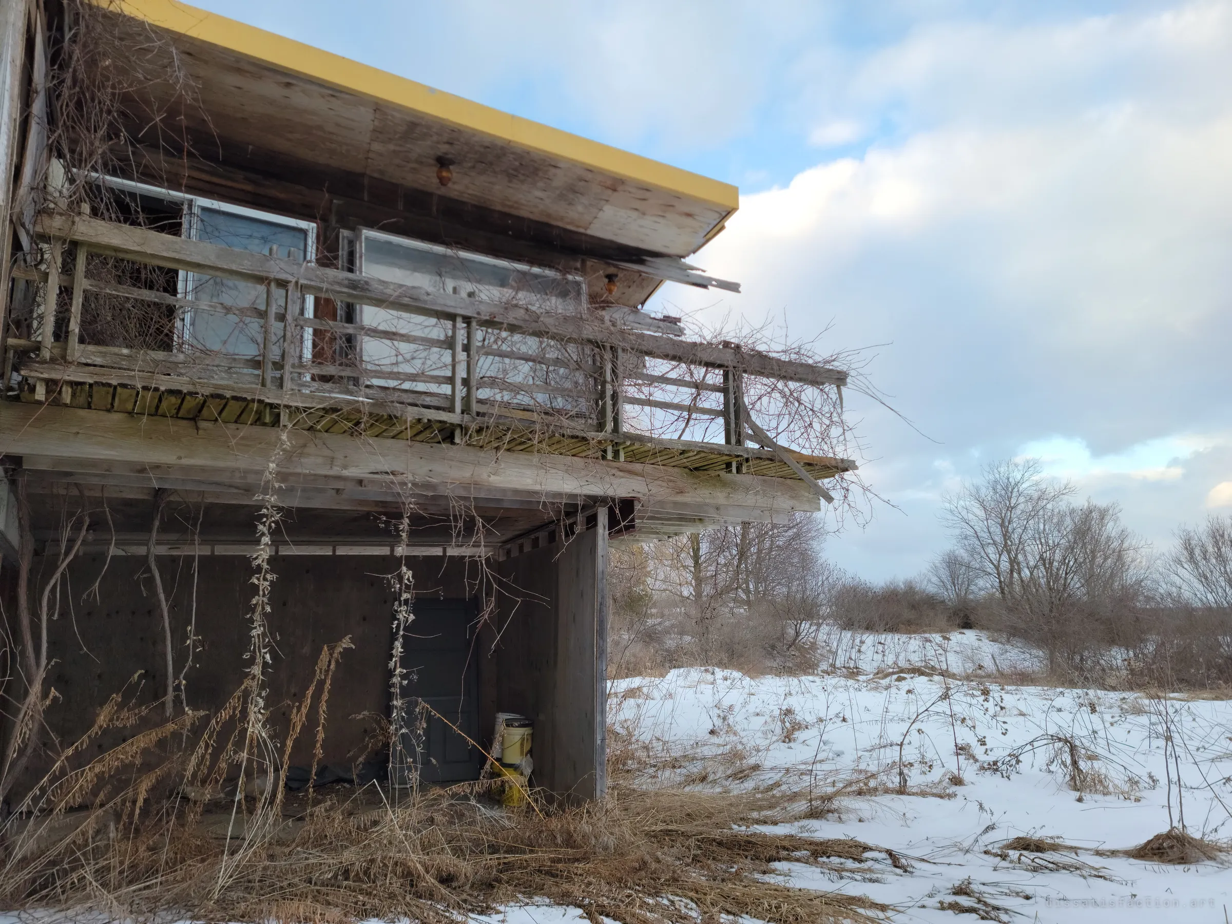 A sagging wooden balcony wrapped in dead vines juts out beneath the yellow roof edge, over a sheltered doorway and tall dry grass. Snowy scrubland stretches away to the right.