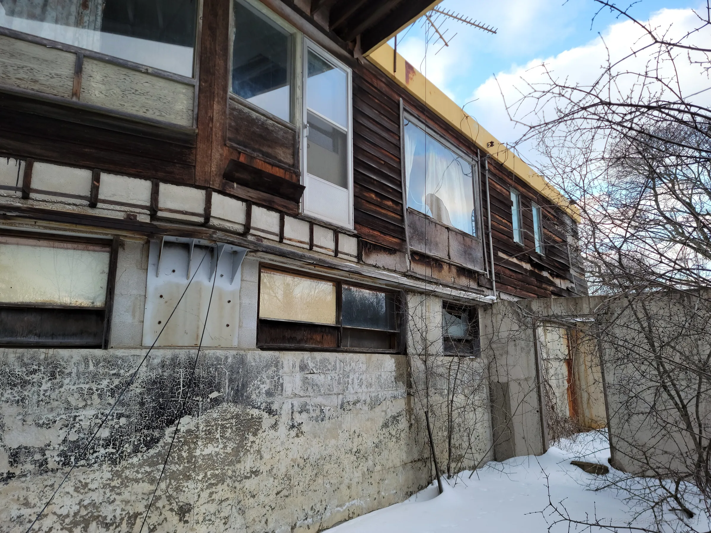 Looking up along the weathered facade: cracked, blackened paint on the block foundation, steel brackets, boarded basement windows and the timber upper floor under a yellow roof edge. Layers of patching show years of neglect.
