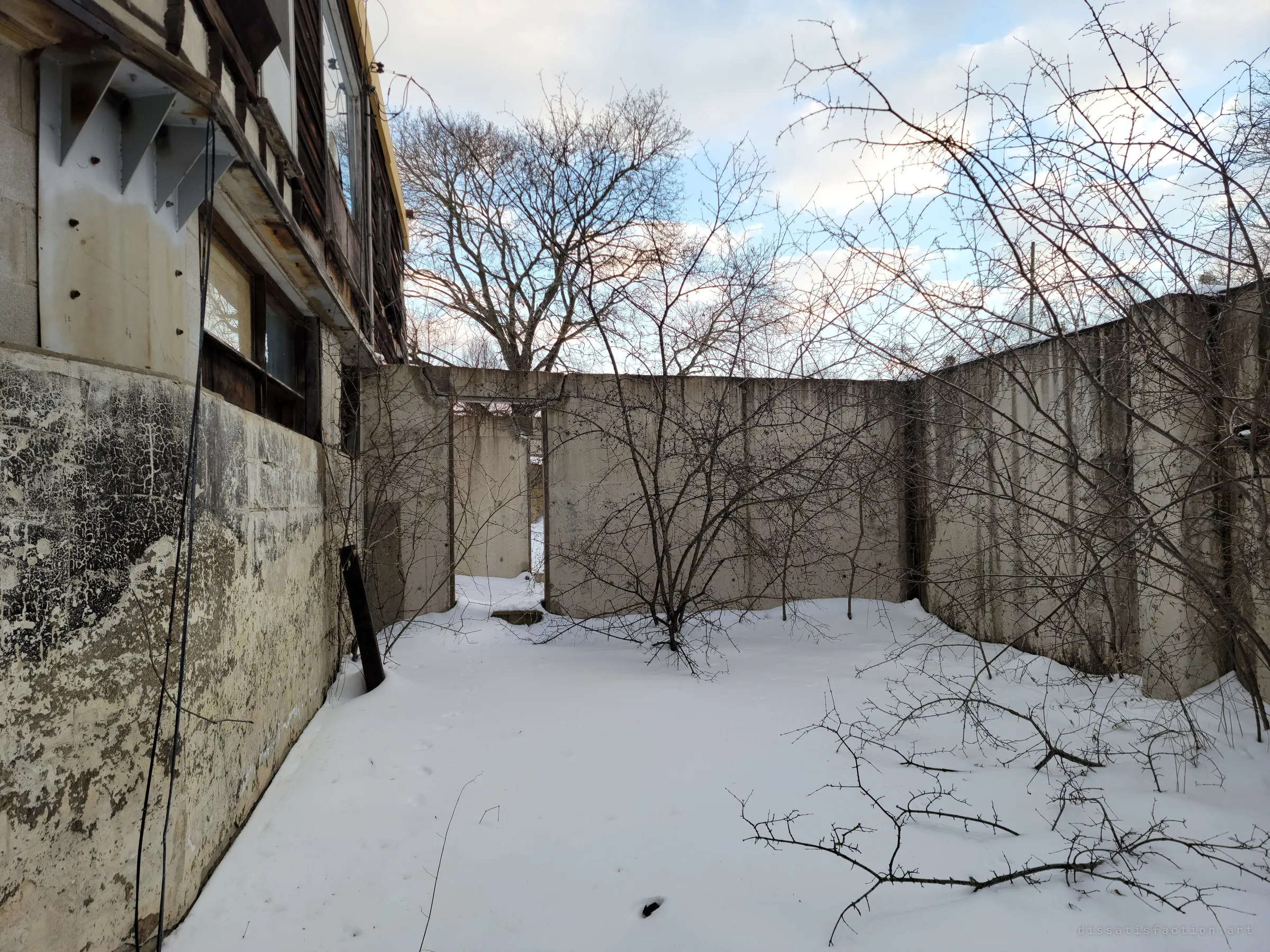 A roofless concrete enclosure filled with snow and bare thorny shrubs, open doorways leading to further empty rooms beside the house. It reads like the foundation of rooms that were never built.