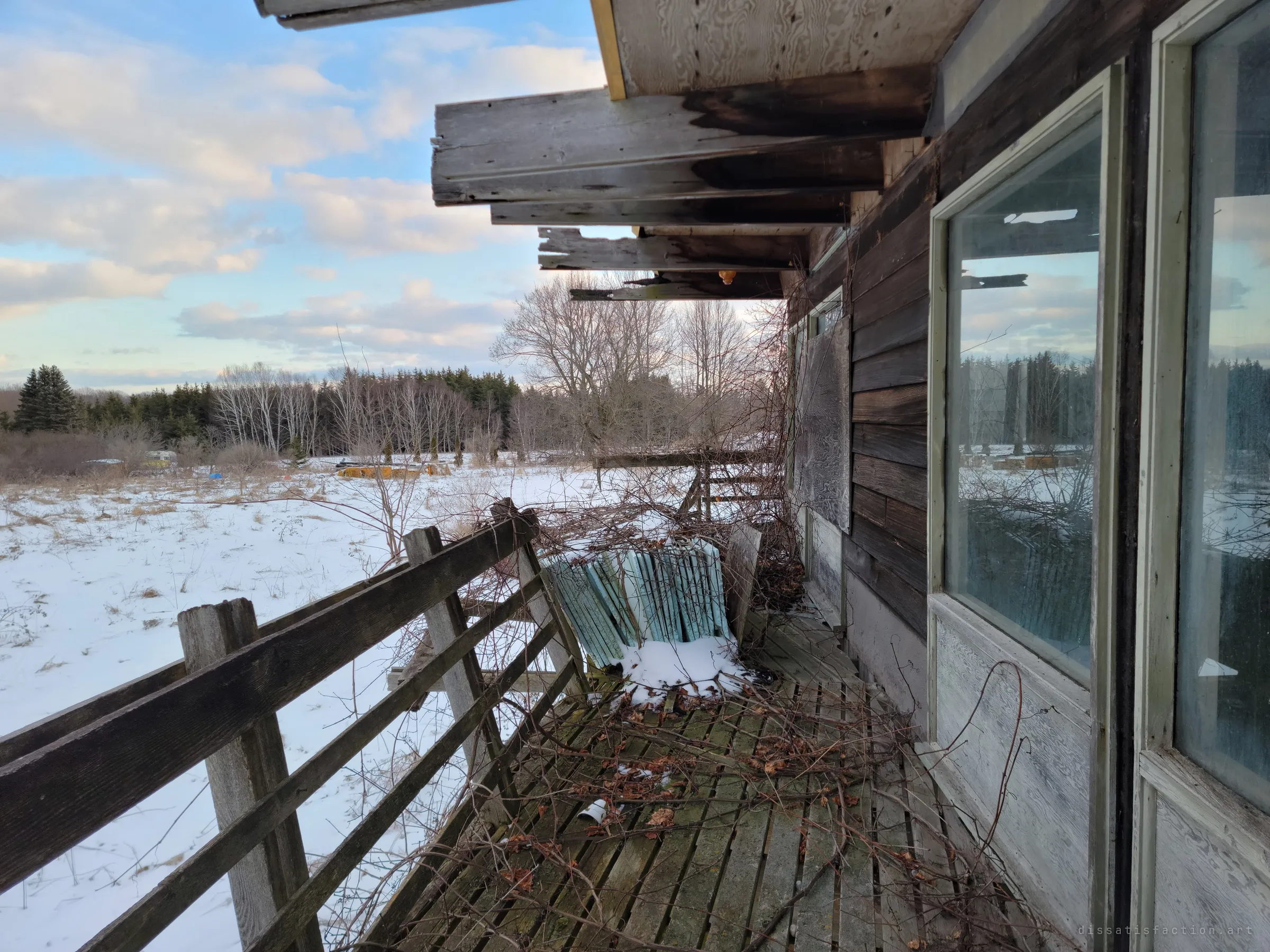 A vine-choked wooden deck runs along the side of the house under broken rafters, a stack of old window screens leaning at the rail, looking out over a snowy field toward a tree line. The windows reflect the winter sky.