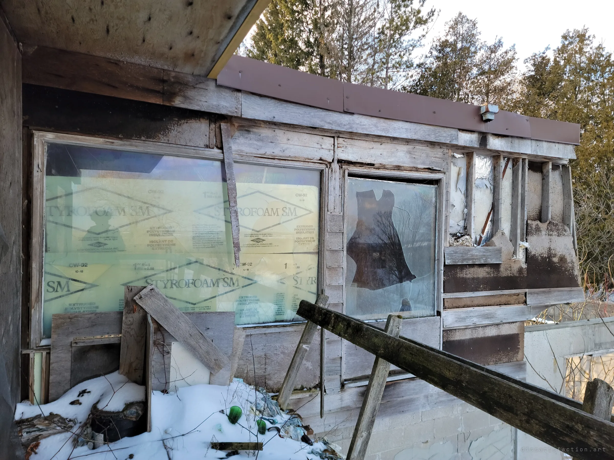 A weathered plank wall with foam insulation boards pressed behind the glass and one pane smashed through, snow and green bottles piled below. The outside of the house is coming apart in layers.