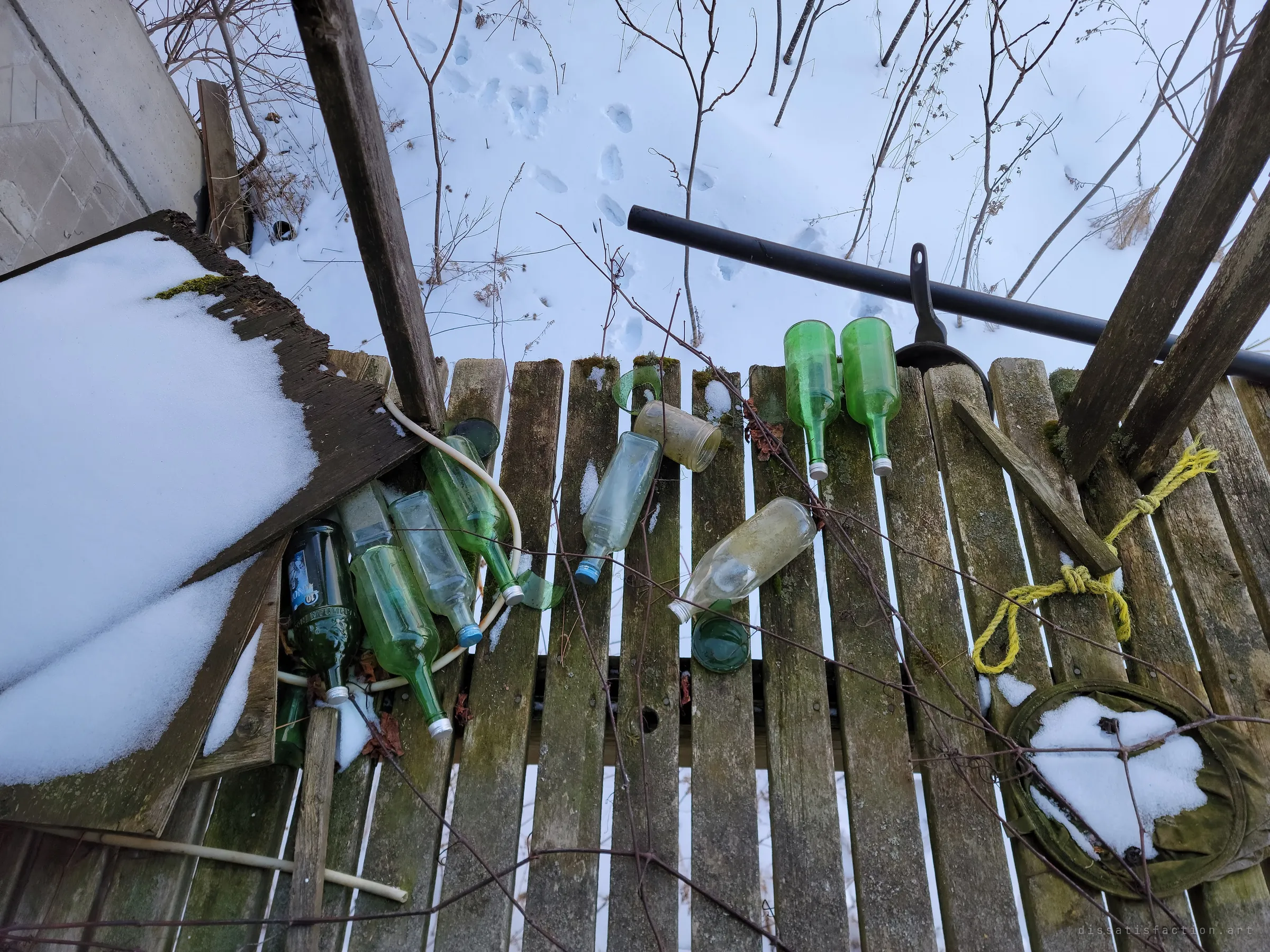 Looking down from a mossy slatted deck at a scatter of green glass bottles, a knotted yellow rope and animal tracks in the snow below. Small leftovers of whoever spent time here.