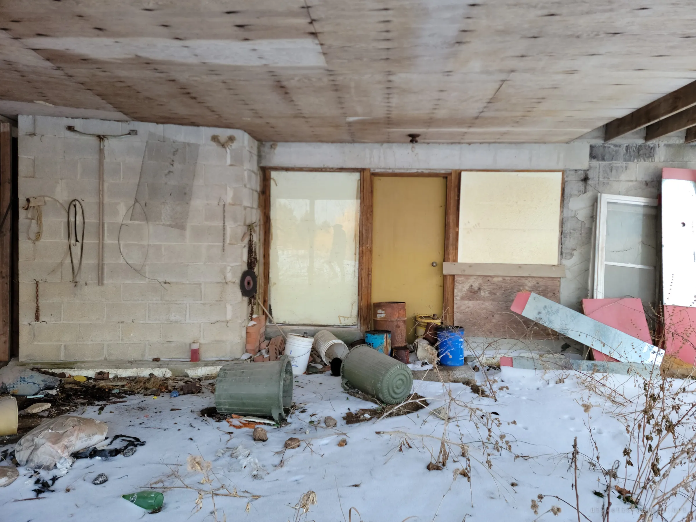 An open carport under a plywood ceiling, a yellow door and boarded windows behind tipped-over bins and paint buckets in the snow. The entrance side of the house, left to the weather.