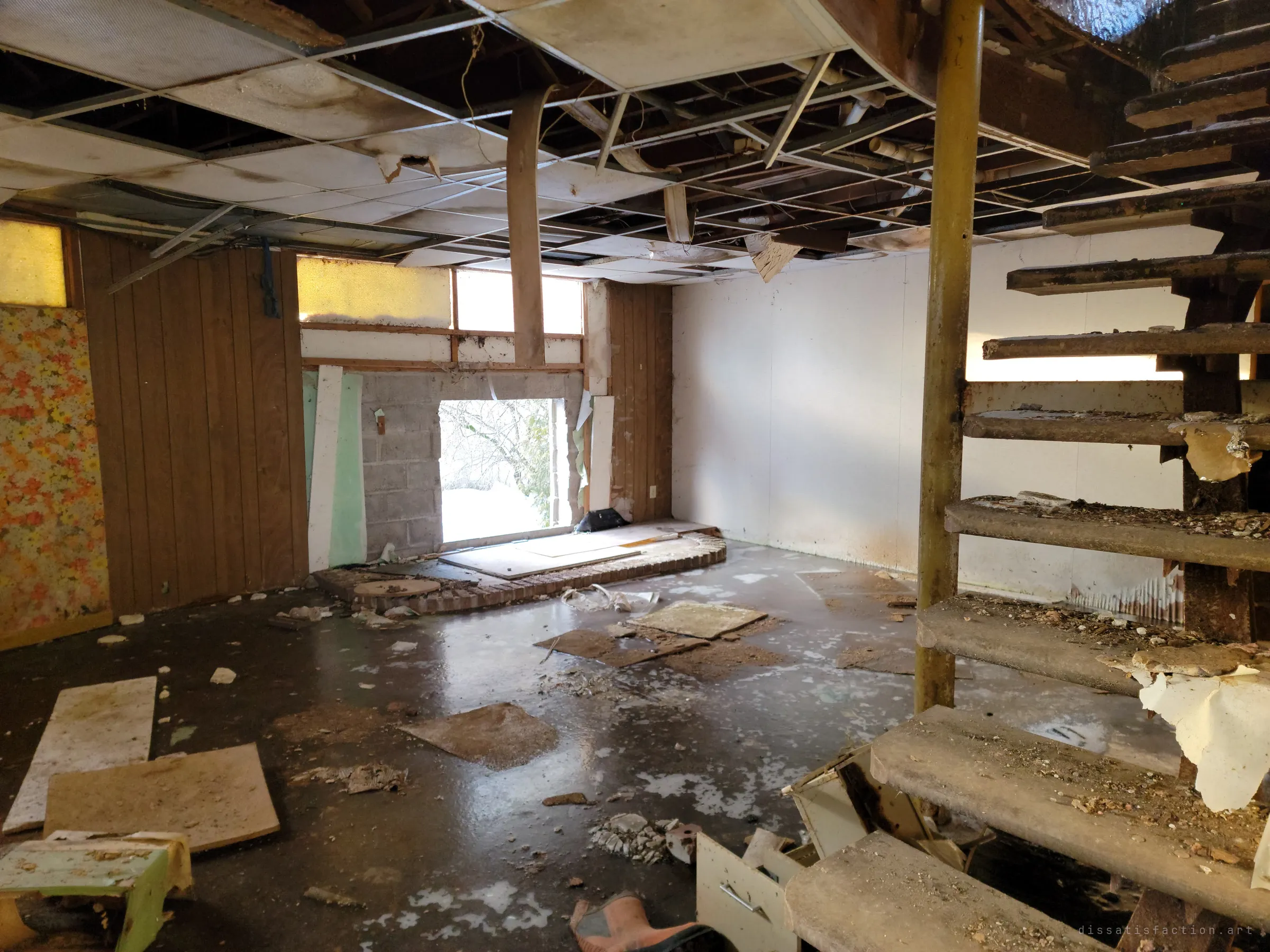 A basement rec room with a collapsing drop ceiling, wood panelling and floral wallpaper, a wall knocked out to the snow and an open-tread stair on the right. The floor is slick with ice and fallen tiles.