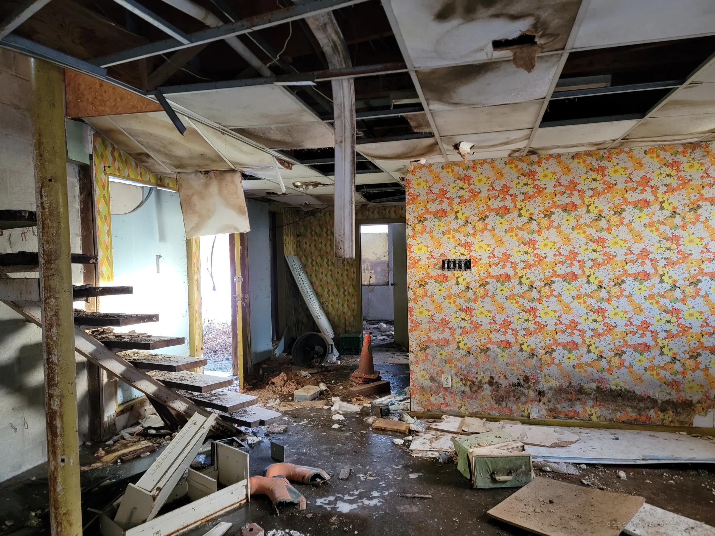 The same basement from the stairs: loud orange floral wallpaper, harlequin-patterned walls, a traffic cone and rubber boots among the debris. The seventies decor survives the rot around it.