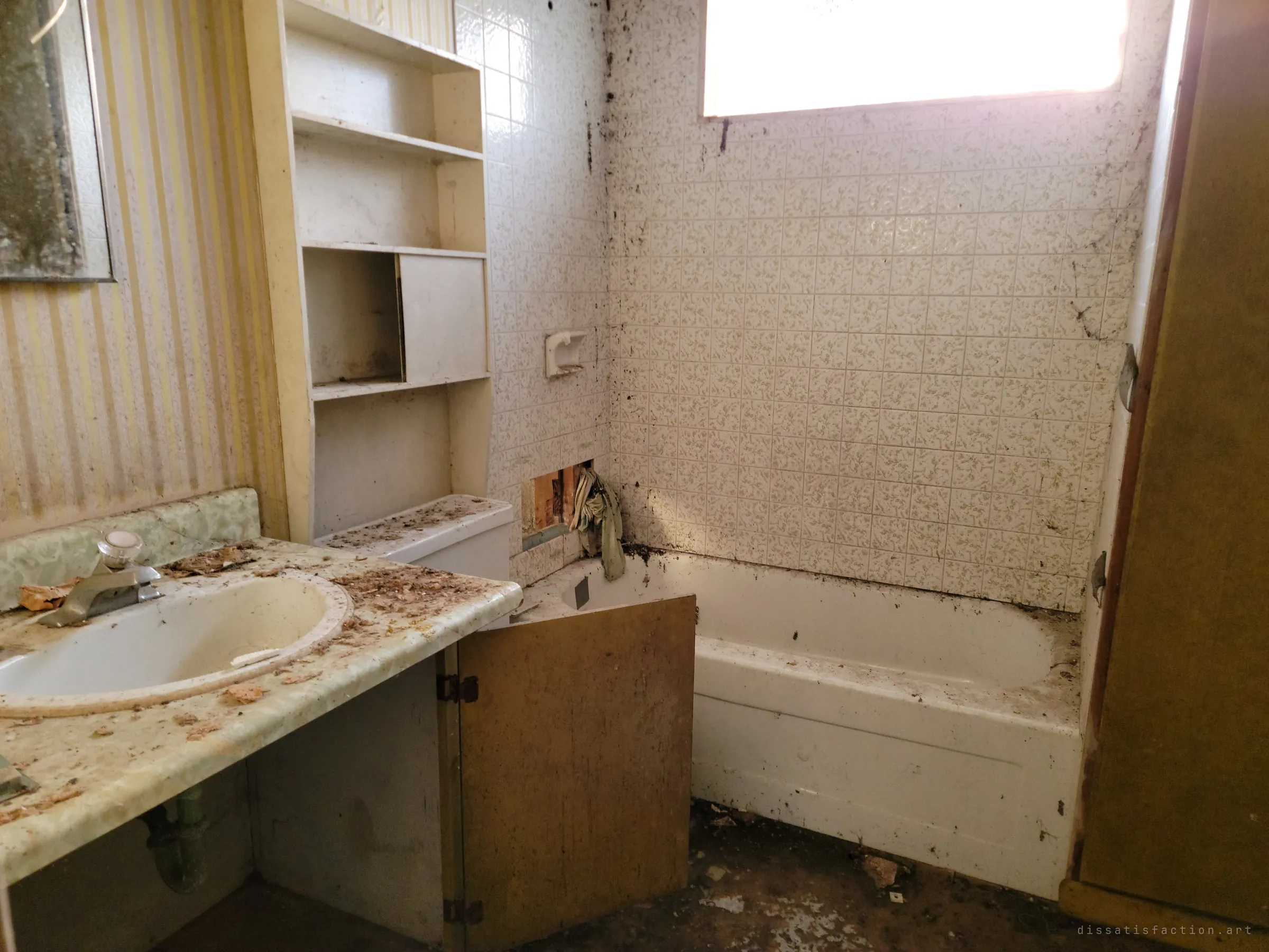 A small bathroom with striped wallpaper, a debris-covered vanity, open shelving and a speckled tiled tub under a bright window. Grime and dirt creep down the tiles.