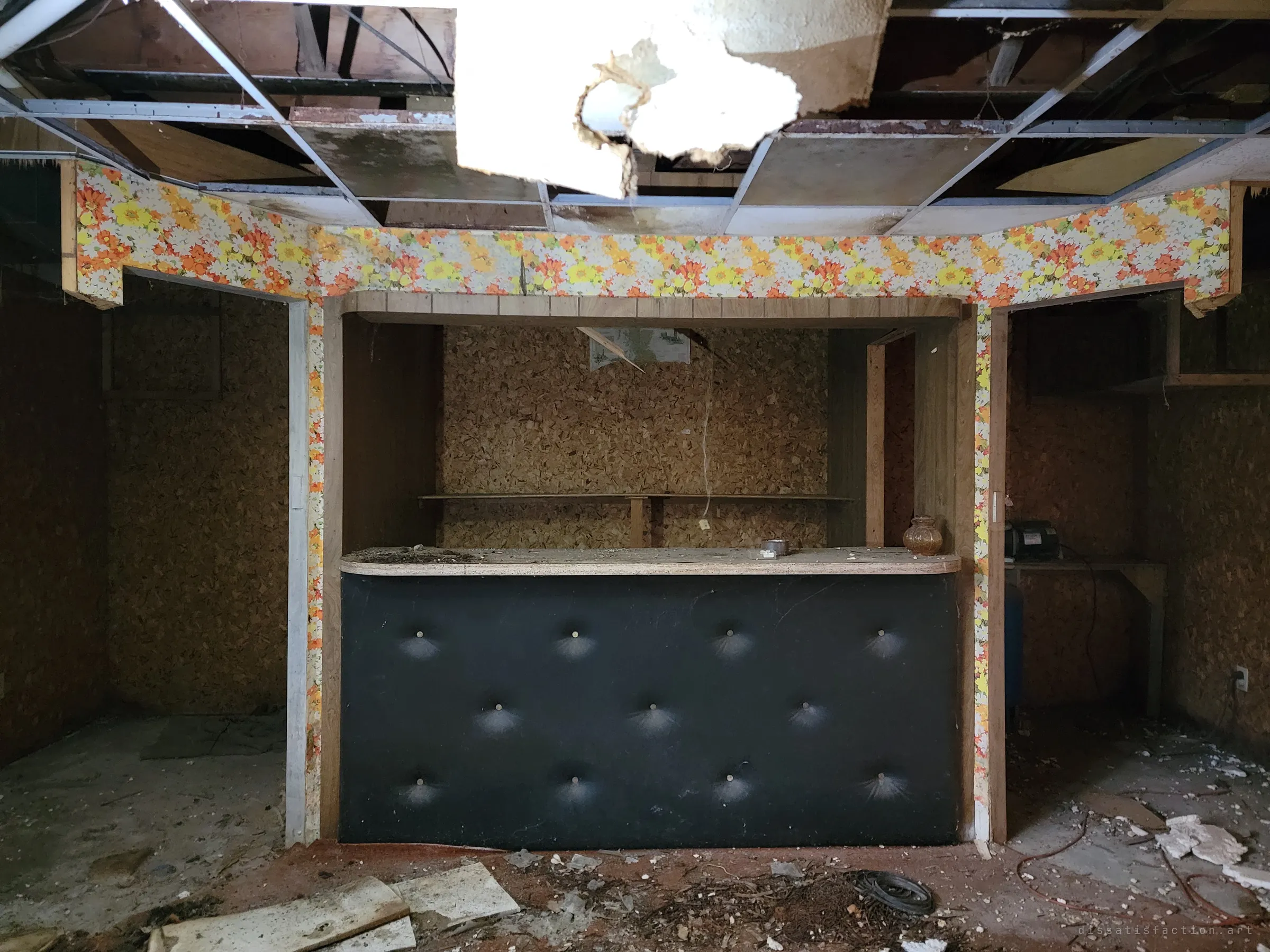 A basement bar framed in orange floral wallpaper, its black tufted front facing the camera beneath a torn-open ceiling. The centrepiece of a long-gone rec room.