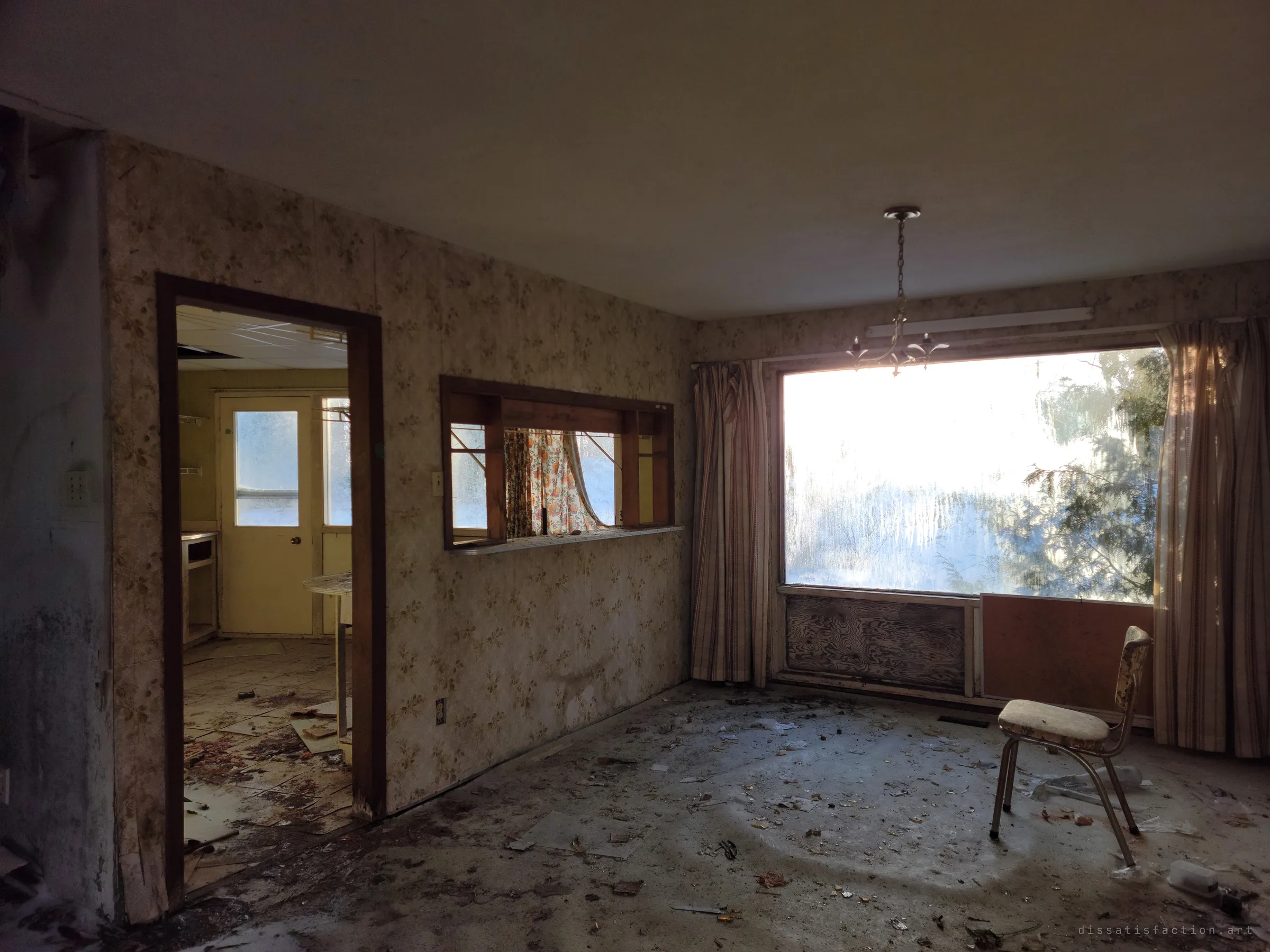 A dining room with faded floral wallpaper, a pass-through to the kitchen, a hanging light and a lone chair in front of a frosted picture window. Winter light falls across the dirty floor.