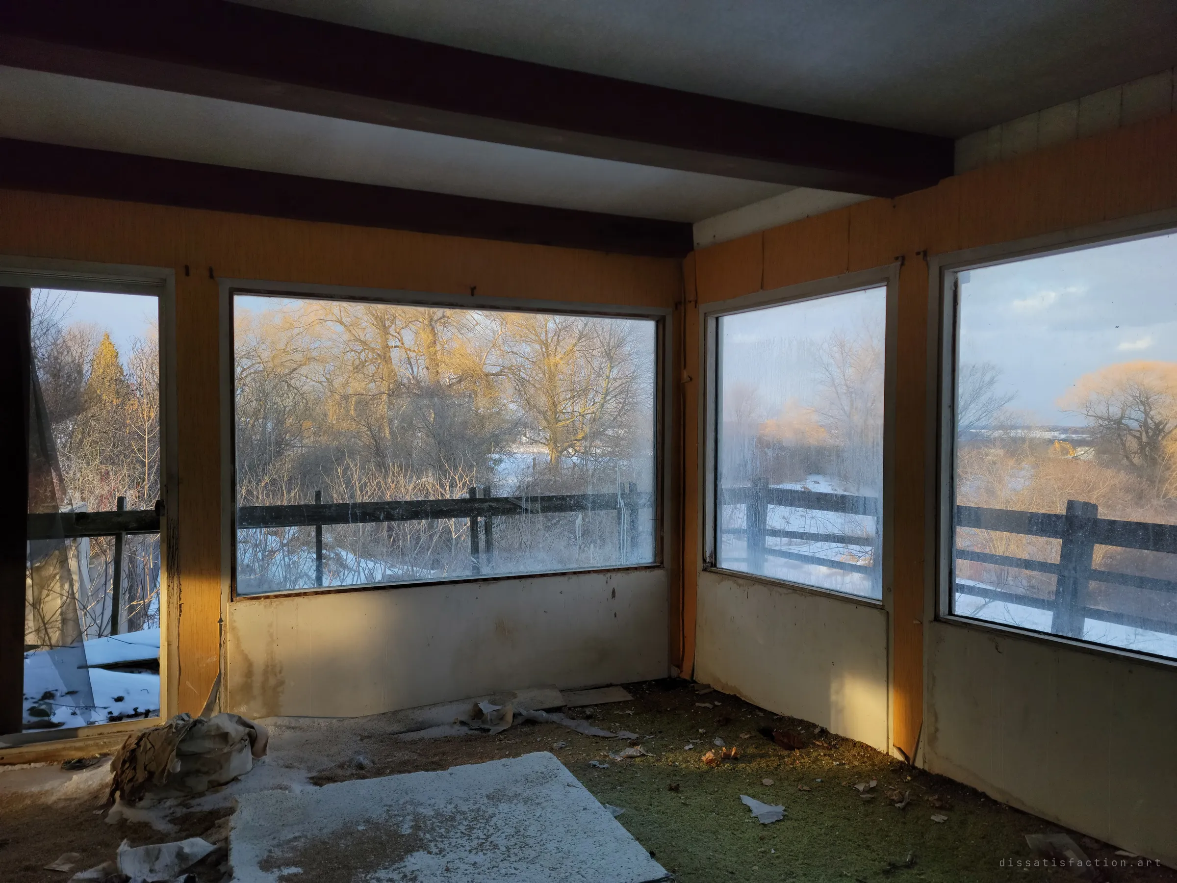 A sunroom with wood-panelled walls and wide windows looking out over a snowy wooden railing and bare trees lit gold by low sun. Snow has blown in across the carpet, showing how open the house is to the weather.
