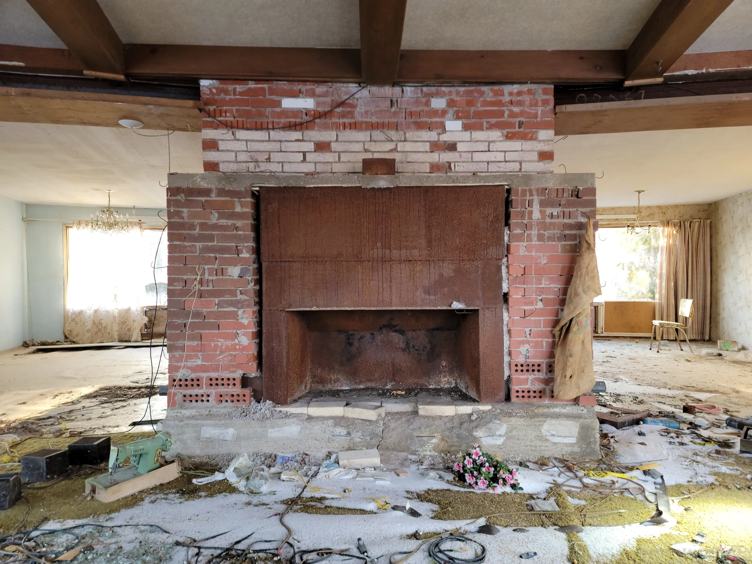 A stripped double-sided brick fireplace stands in the middle of the open living space, its rusted steel firebox bare, with a green sewing machine, cables and a bunch of fake pink flowers on the snow-dusted carpet in front. Chandeliers still hang in the rooms on either side.