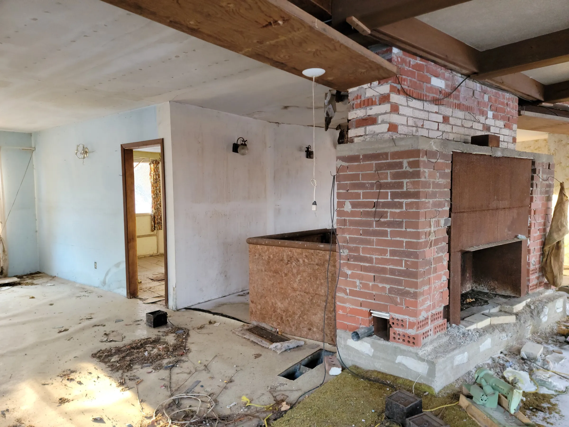 The same brick fireplace seen from the side, beside a low panelled railing and a bare pale-blue room with brass wall sconces and an open door to a flowered-curtain room beyond. A single bare bulb hangs on its cord from the ceiling.