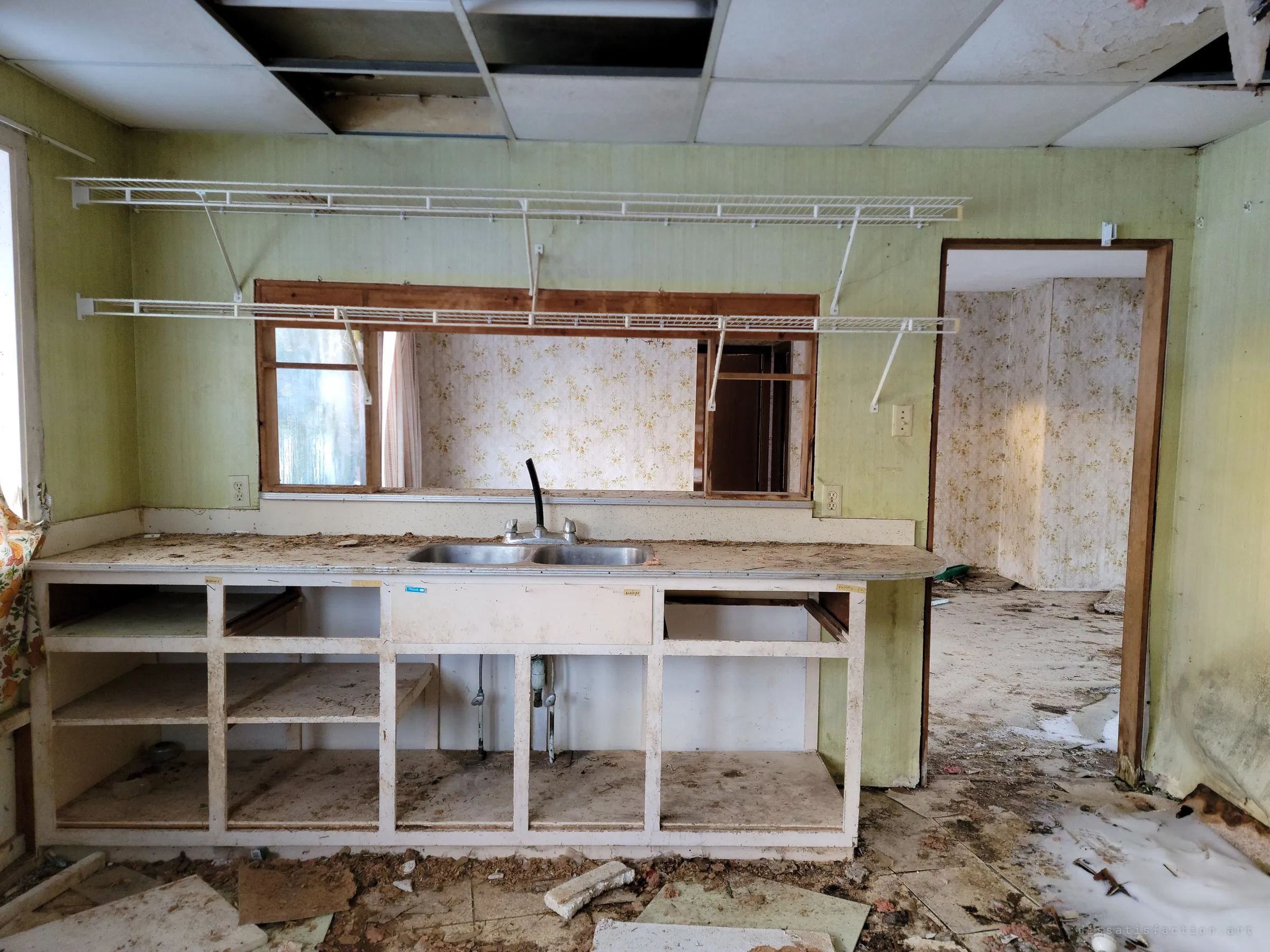 A gutted kitchen with a double steel sink in an empty frame of cabinets, wire shelving above and a pass-through window into a floral-wallpapered room. Small handwritten labels are still taped to the cabinet fronts.