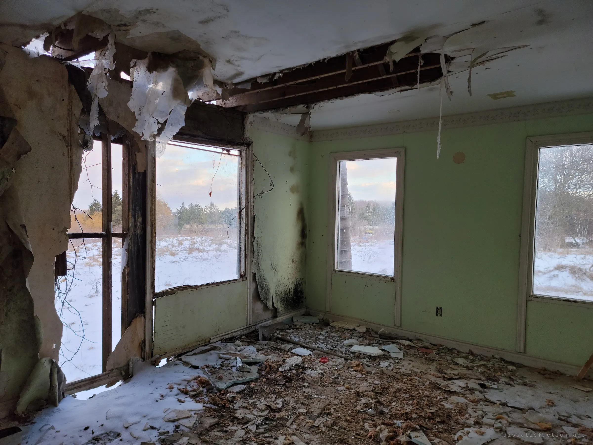 A mint-green bedroom where the ceiling has torn open and the wall has rotted through to the studs, with snow drifting in over the debris-covered floor. Three windows look out onto a white, empty field.