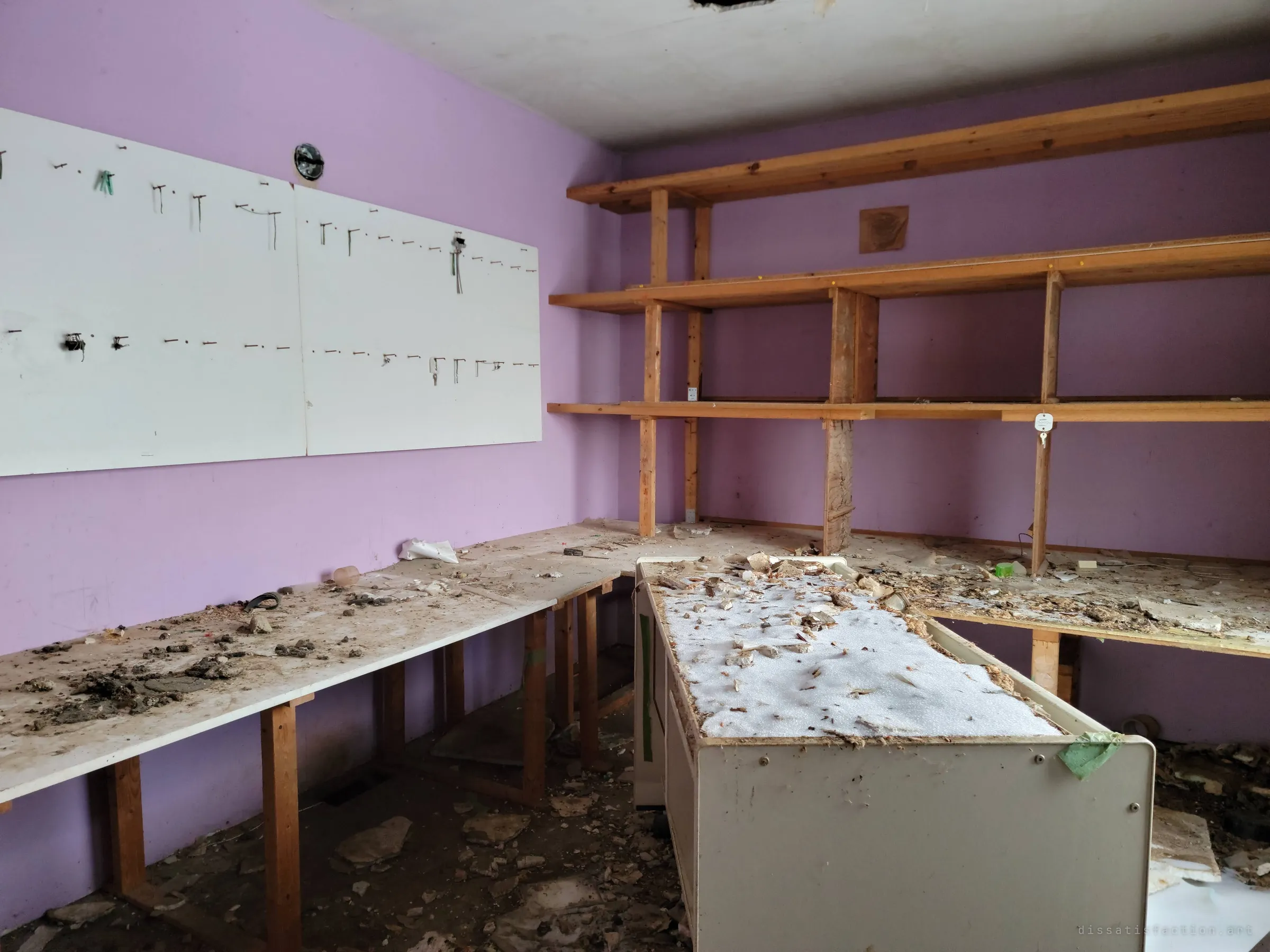 A lavender-walled workroom with long plywood benches, empty wooden shelving and a white pegboard where rows of hooks once held keys or tools. Plaster and debris cover every surface.