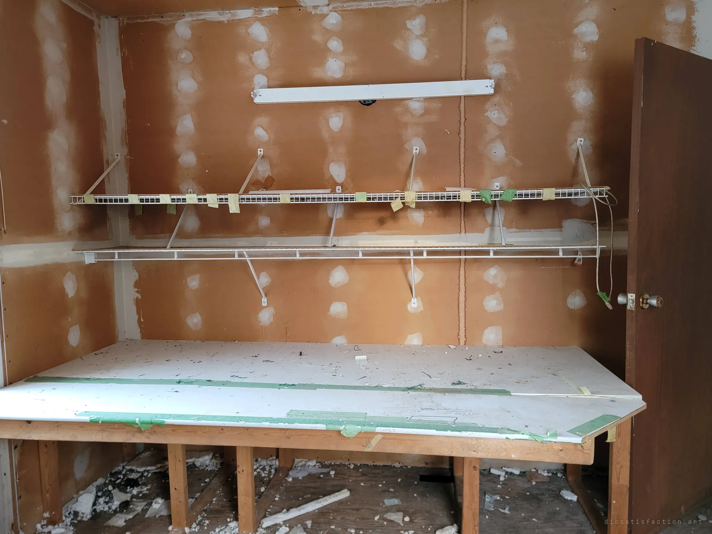 A small room with patched drywall, two wire shelves covered in masking-tape labels and a large white worktable edged with green tape and faint pencil notes. A dark wood door stands open at the right.