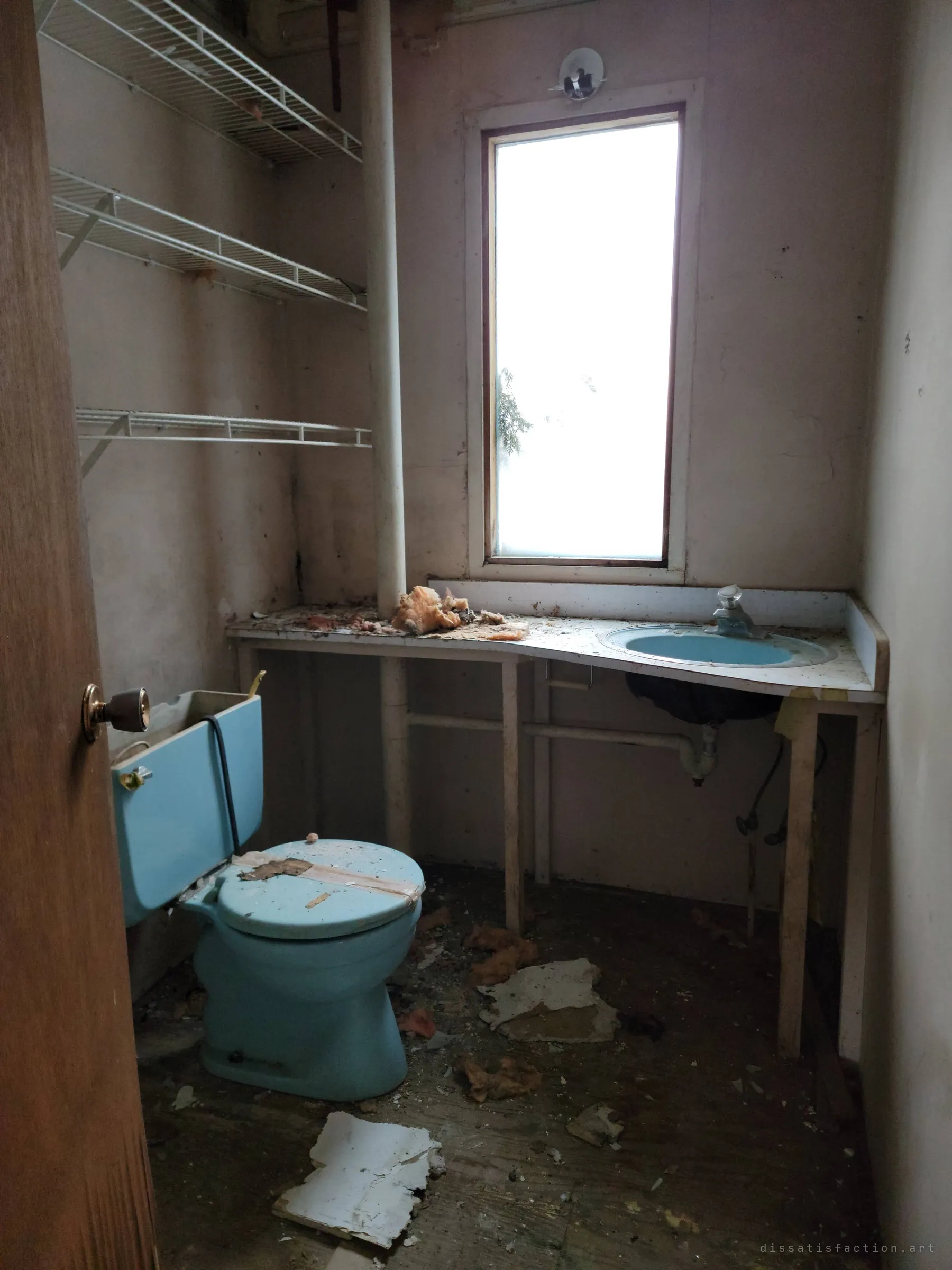 A narrow powder room with a pale-blue toilet and matching sink set into a long counter, lit by a single tall window. Insulation and ceiling fragments lie on the counter and floor.