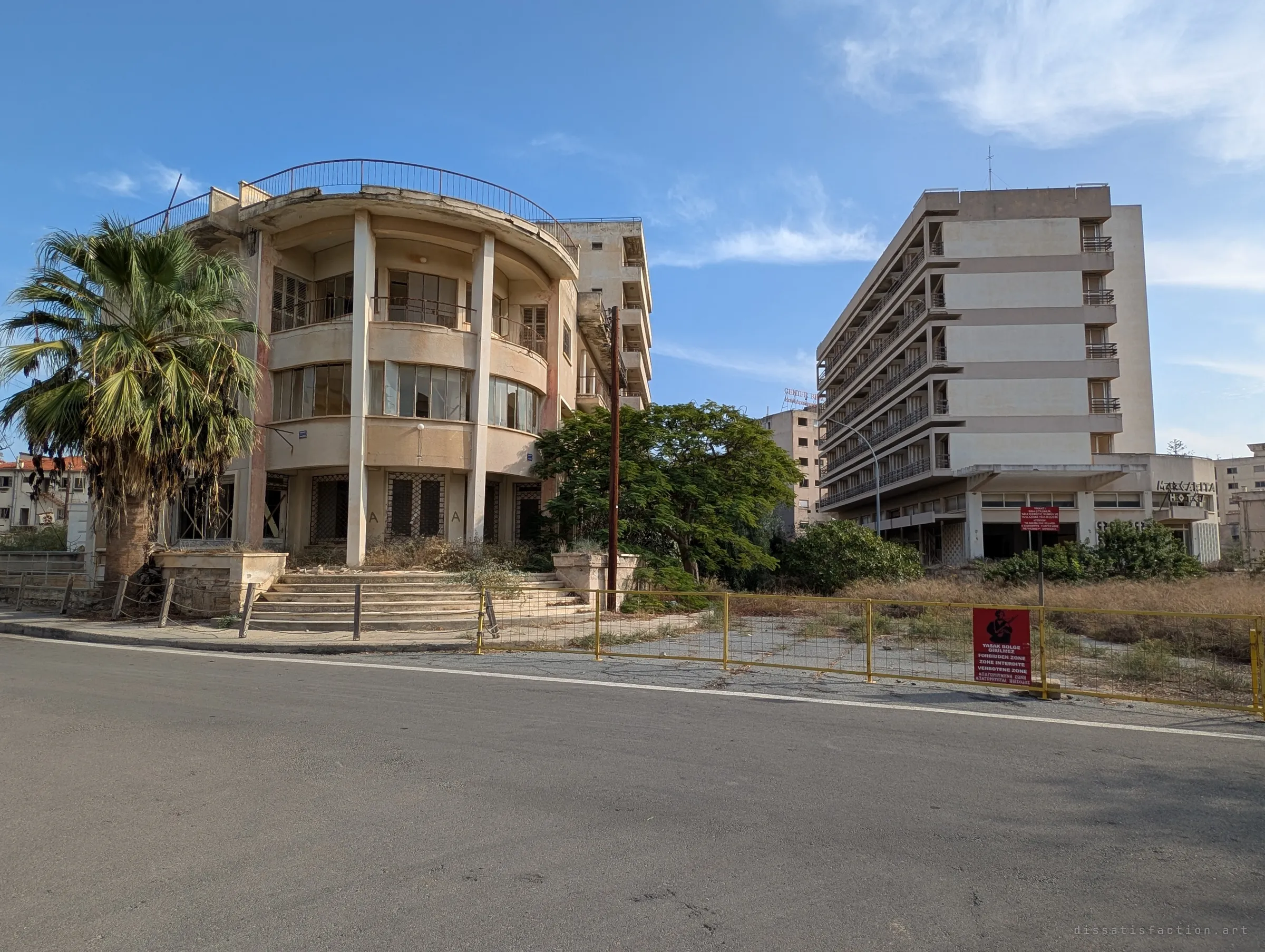 A curved-front villa and a tall balconied hotel stand empty behind a yellow barrier fence and a red forbidden-zone sign. The empty road and overgrowth show a resort frozen in time.