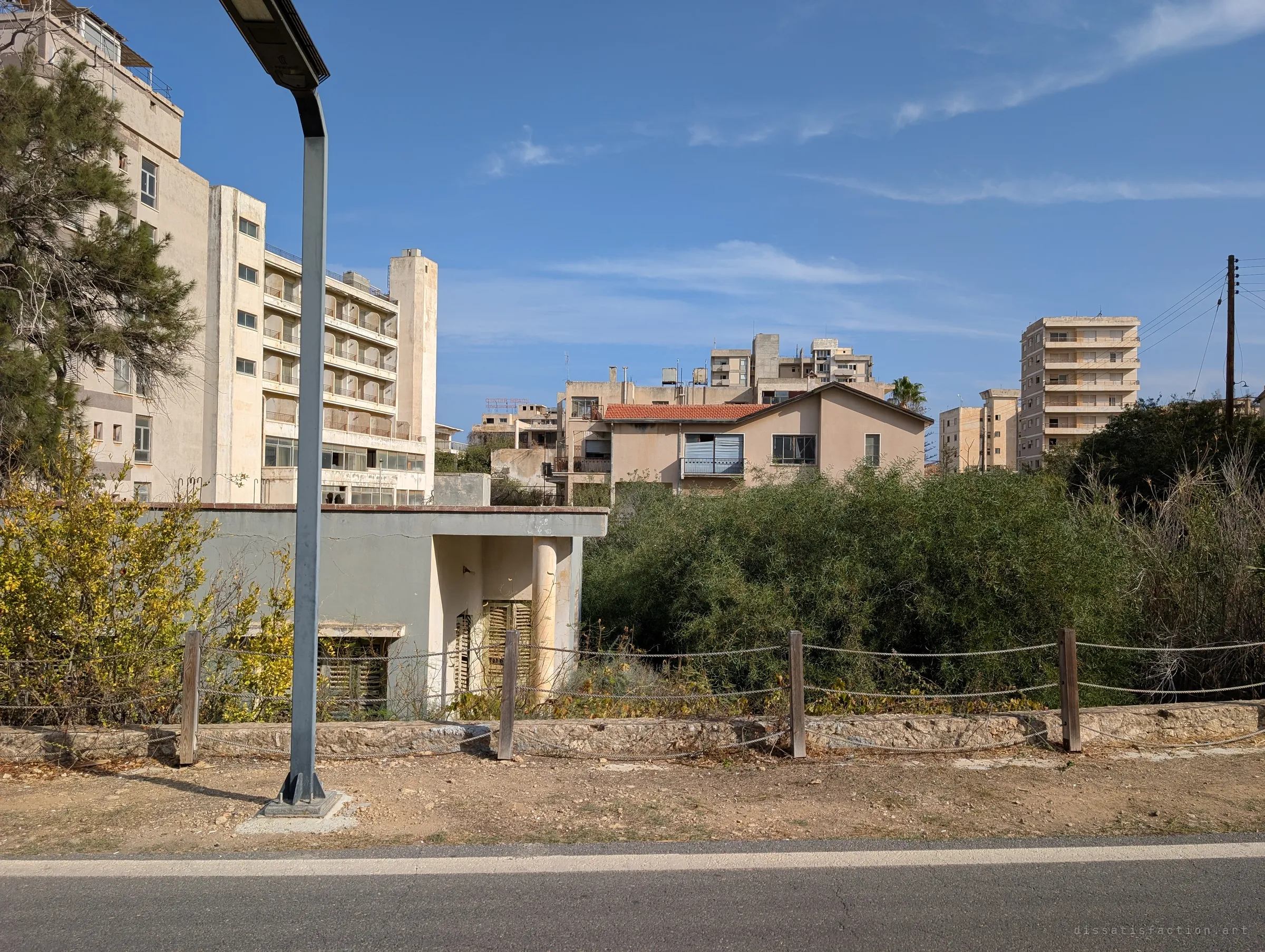 Behind a rope fence and dense scrub, tiled-roof houses and hollow hotel towers rise against a clear sky. Nothing moves; the street lamp is the only new thing in frame.