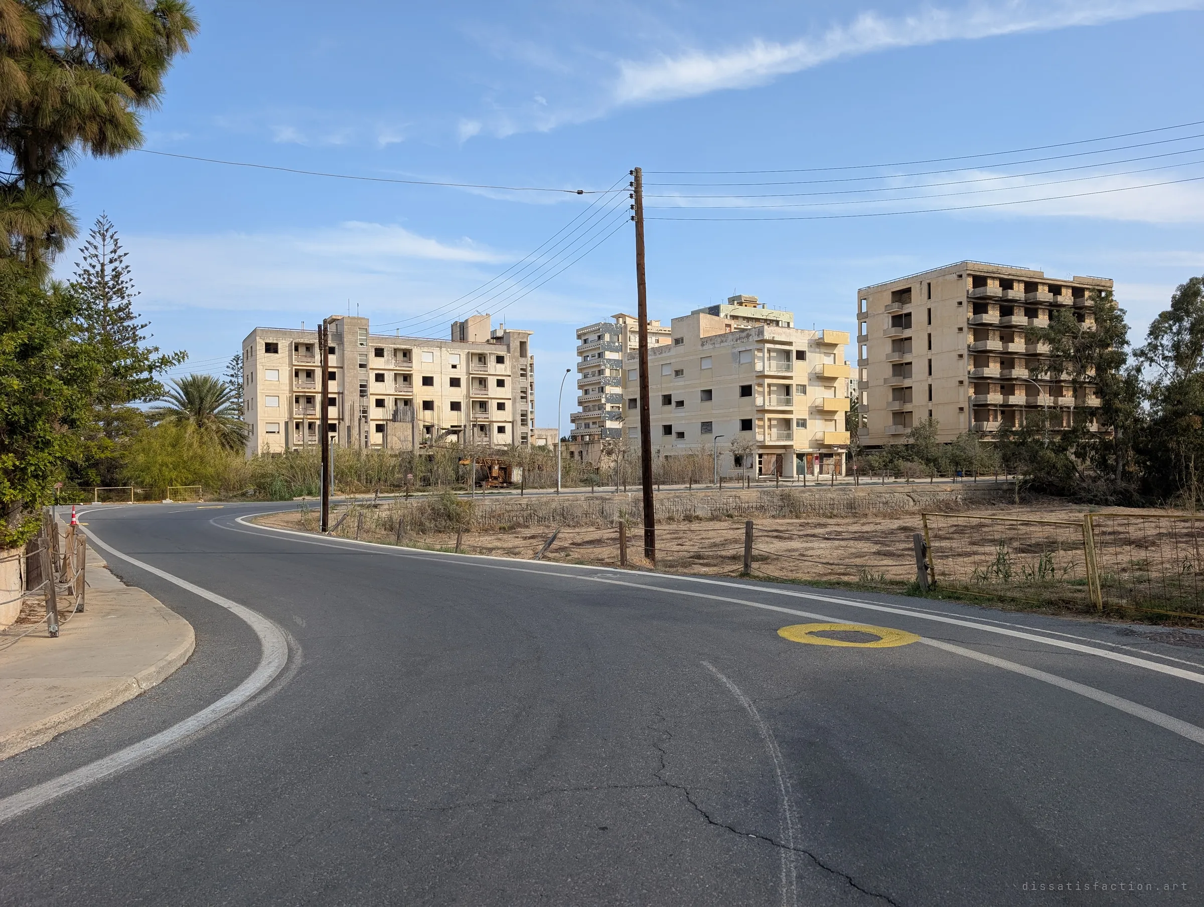 A curving empty road leads past a row of gutted apartment blocks with blank window holes. Power lines and a painted yellow road circle are the only signs of upkeep.