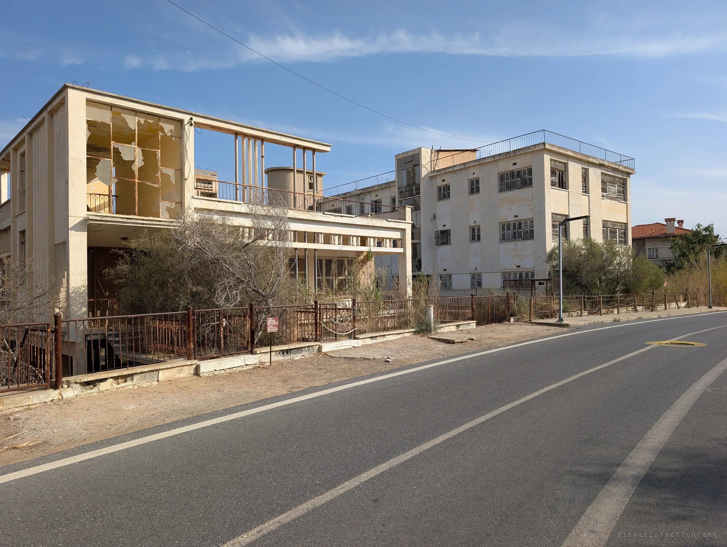 A modernist villa with a shattered glass upper room and a shuttered three-storey block sit behind rusted railings along an empty road. Decades of neglect show in every broken pane.