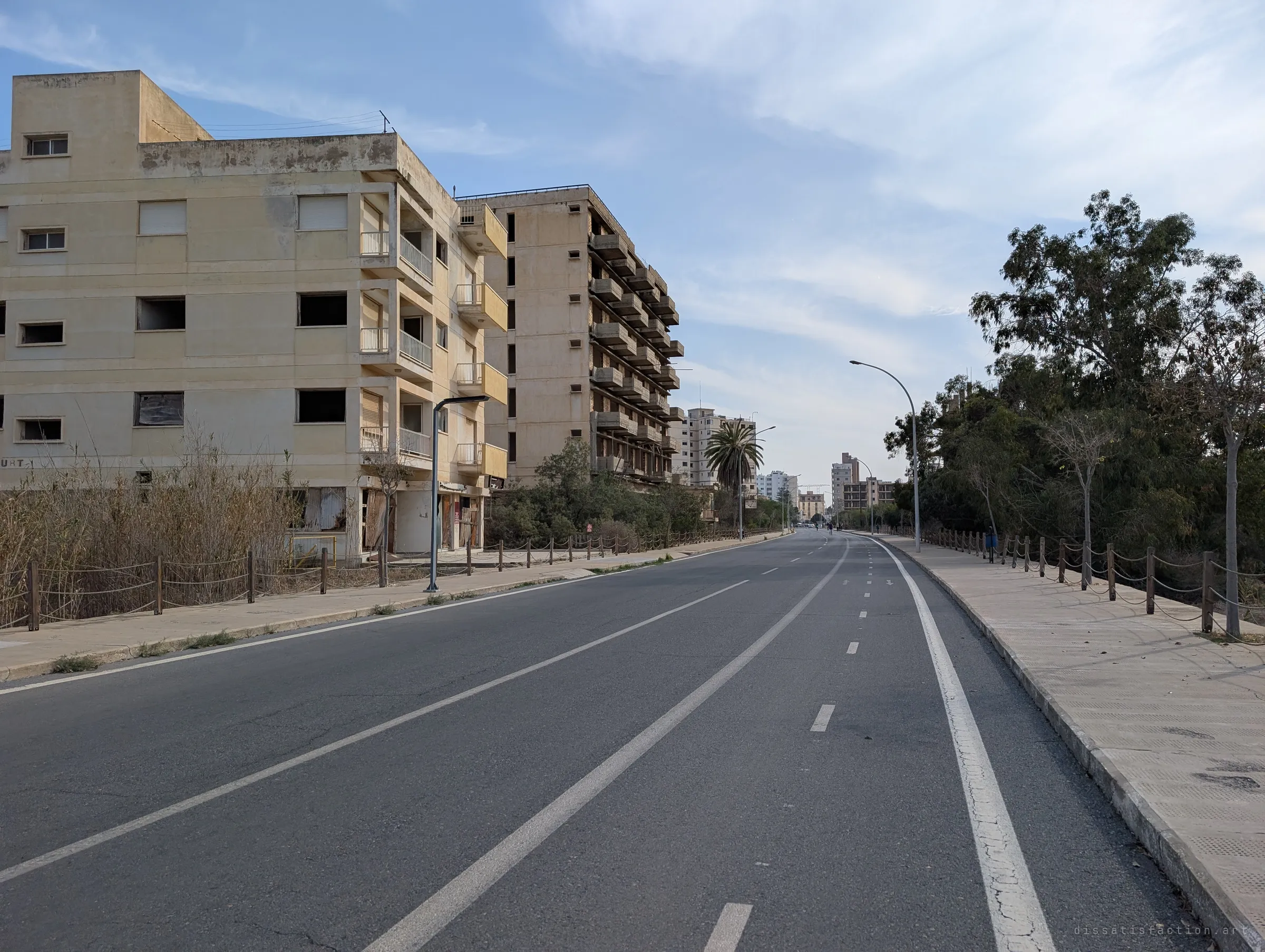 A wide, freshly marked boulevard runs past empty balconied hotels toward a distant skyline. The new asphalt contrasts sharply with the abandoned buildings beside it.