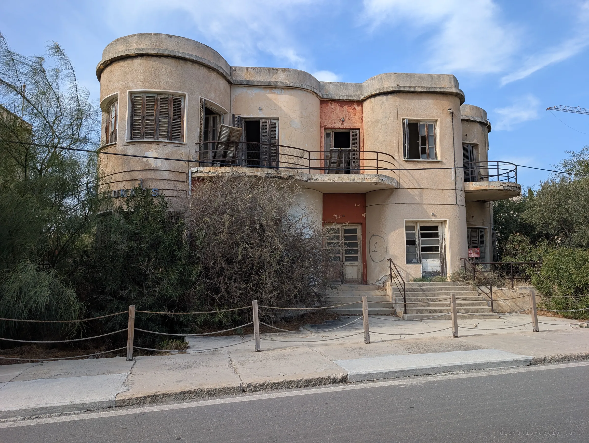 An art-deco house with rounded bays, broken shutters and a painted number on its wall hides behind overgrown shrubs. Its curved balconies still hold their original railings.