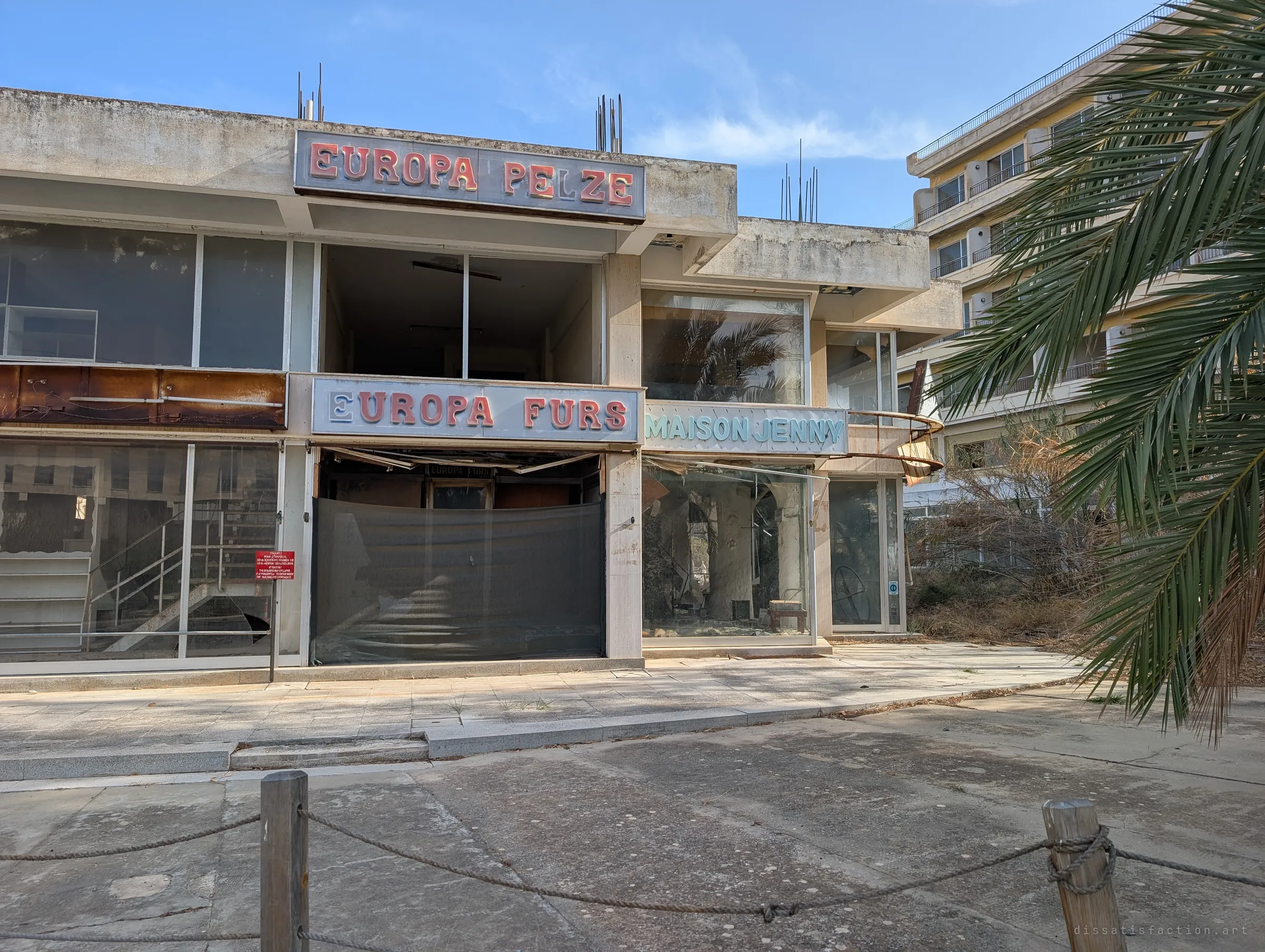 A row of abandoned boutiques with faded retro lettering, broken glass and a sagging shutter. Their signs, frozen for decades, show the district's former glamour.